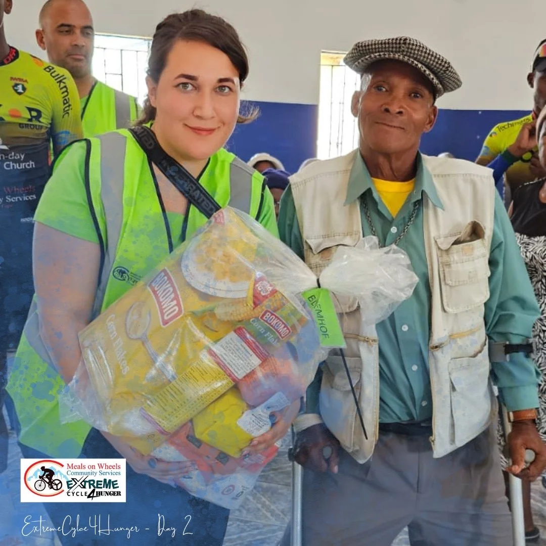 A woman in a yellow safety vest holding a large bag of groceries, standing next to an older man with a cane, wearing a patterned hat, a light vest, and a blue shirt. They are inside a community service setting with other people in the background.