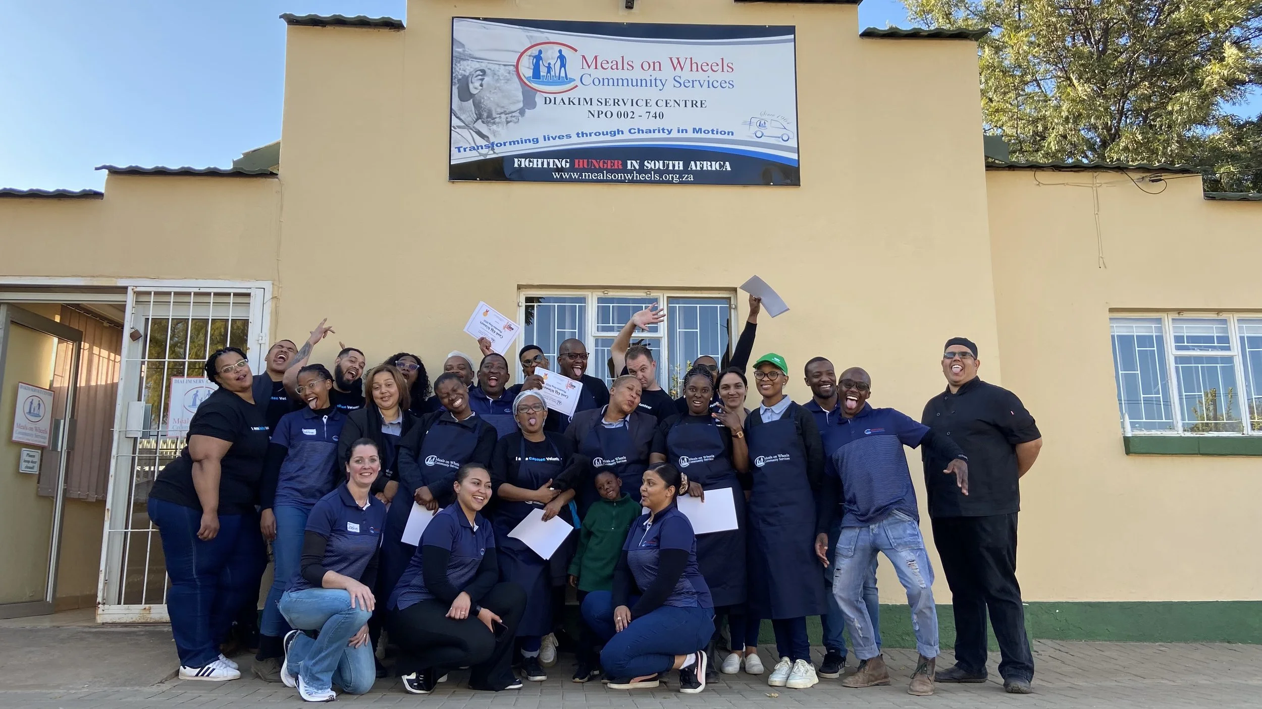 Group of people standing in front of a building with a sign that reads "Meals on Wheels Community Services." The group is smiling and making various gestures, some holding papers or certificates. Training in the Northern Cape