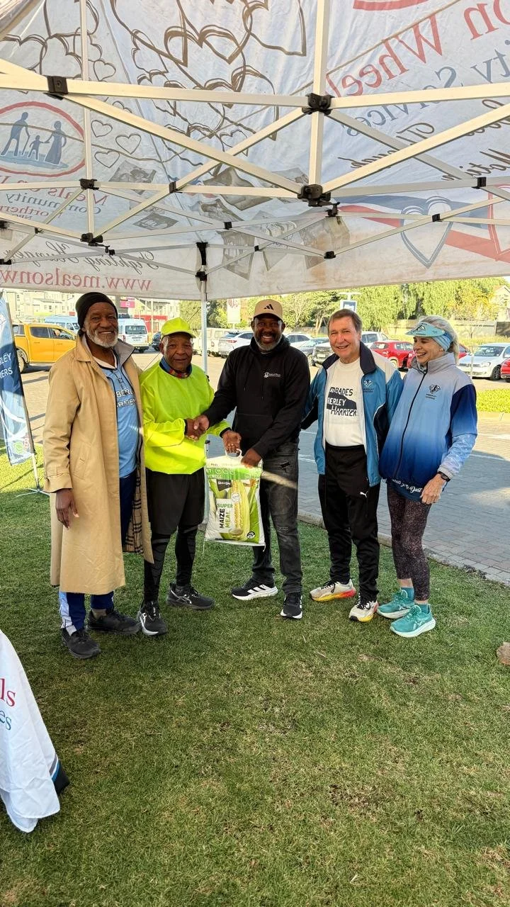 Five people standing under a canopy, shaking hands and smiling. They are outdoors on grass, with a parking lot and trees in the background. One person is holding a bag, and some are wearing athletic clothing. Running for a hunger free South Africa