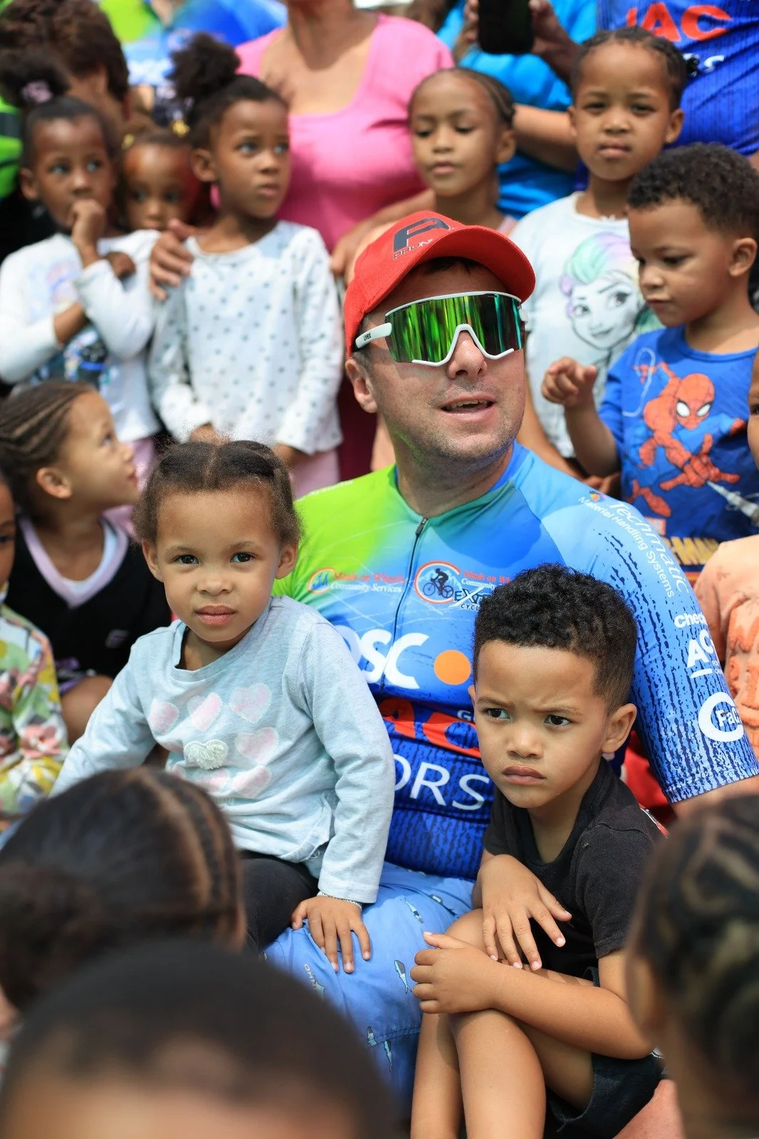 A man with sports sunglasses and a cycling jersey is surrounded by young children of various ages and ethnicities, some sitting on his lap and others standing around him. The scene appears to be outdoors with a large group visibly gathered.