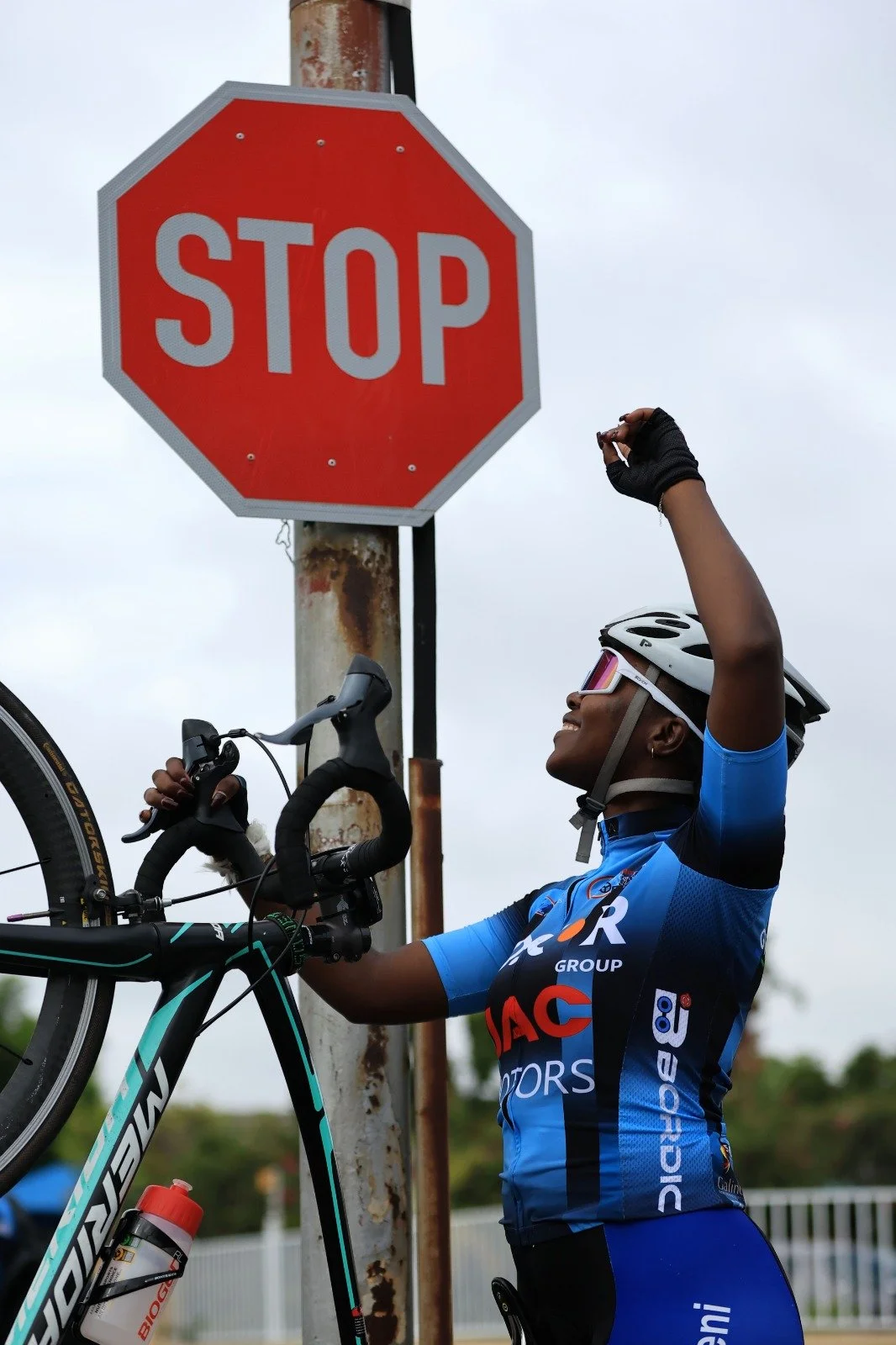 A woman in cycling gear, wearing a helmet and sunglasses, is holding her bike and adjusting a stop sign.