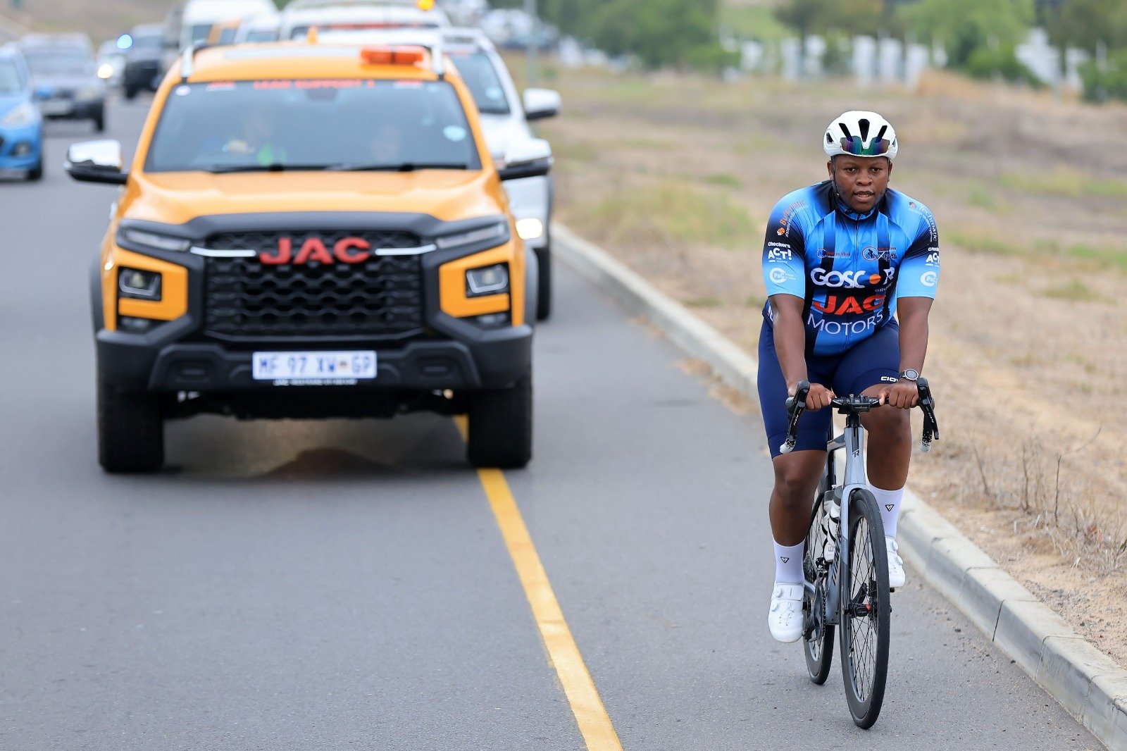 A person wearing a blue cycling jersey and a white helmet riding a black bicycle on the side of the road, with a yellow safety line, and a row of cars including an orange JAC vehicle ahead on the road.