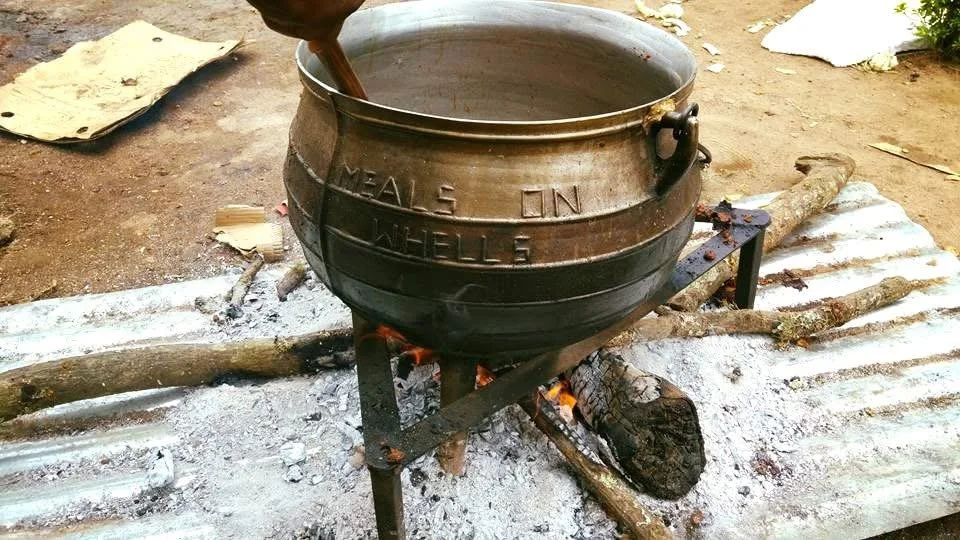 A large metal pot over an open fire with sticks and wood to support it, cooking outdoors on a bed of white ashes. Meals on Wheels in Rural South Africa