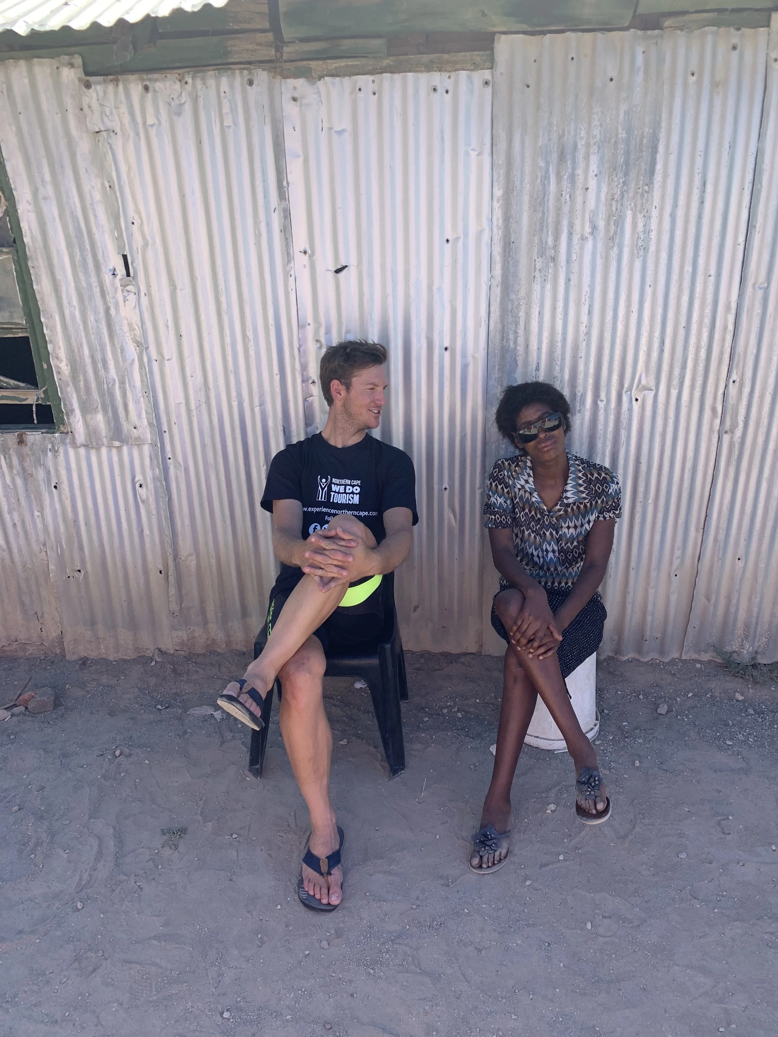 Two people sitting against a corrugated metal wall outdoors. The person on the left is a light-skinned man with short brown hair, wearing a black T-shirt and sandals, who is smiling and looking at the woman. The person on the right is a dark-skinned woman with short black hair, wearing sunglasses, a patterned shirt, a skirt, and sandals, sitting on a white bucket with her hands on her lap.