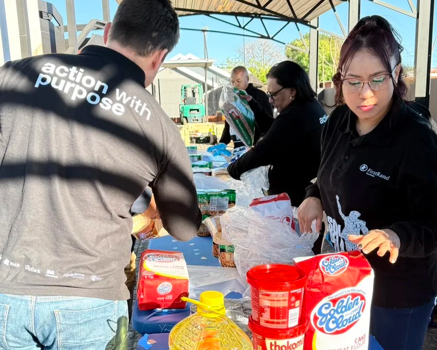 People working together at a food distribution table outdoors, sorting and packing food items, with various food packages and supplies on the table.