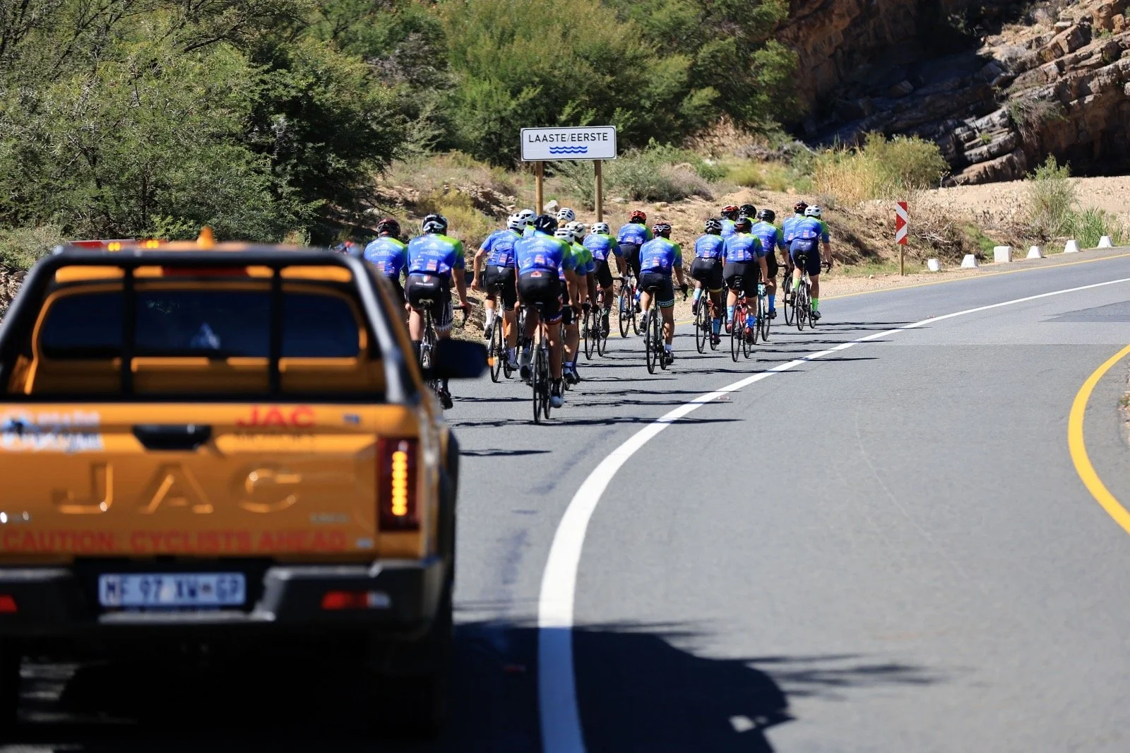 A group of cyclists riding on a winding mountain road, closely packed, with a yellow vehicle in the foreground and trees on both sides of the road.
