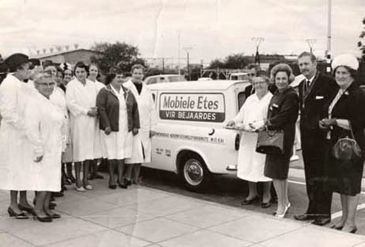 Group of women and men standing outside with a small vehicle that has a sign reading 'Mobile Etes' and 'VIR BEJAARDES' on the side. The Start of Meals on Wheels Community Services in 1964