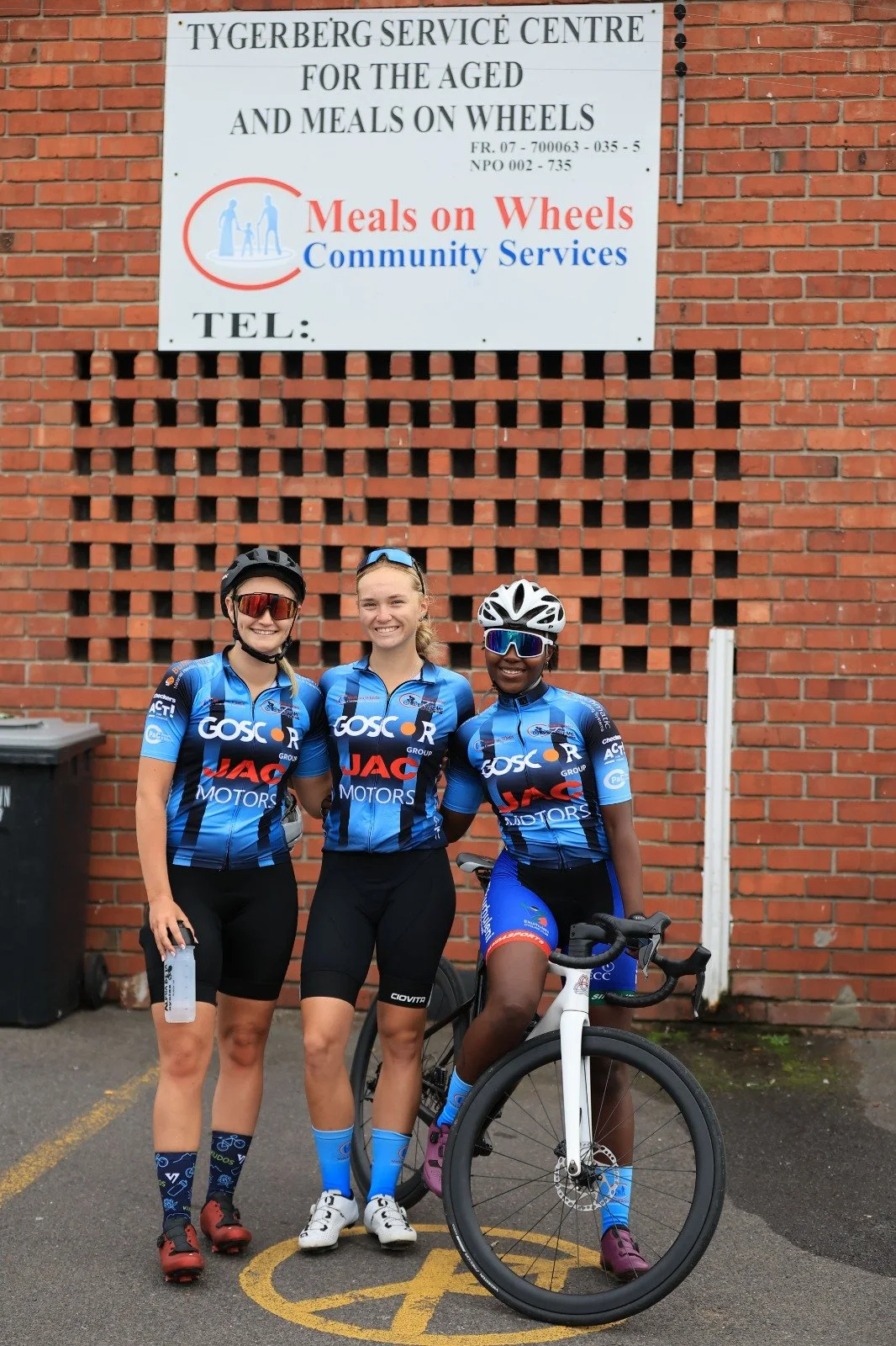 Three female cyclists standing in front of a brick wall, wearing blue cycling jerseys with sponsors' logos, helmets, and sunglasses. One woman is holding a water bottle, and another is sitting on a white bicycle.