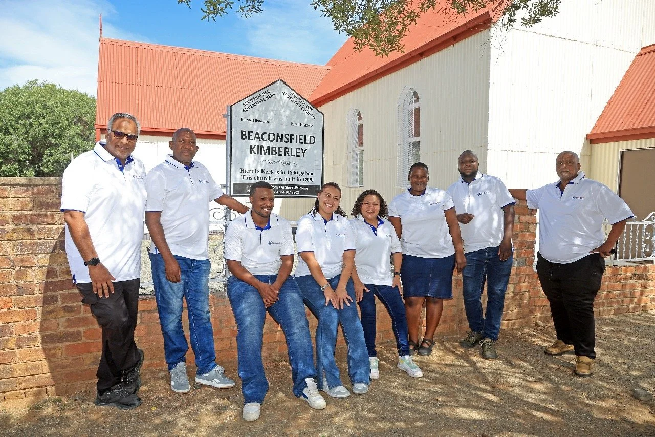 Group of nine people standing outside near a church sign that reads 'BEACONSFIELD KIMBERLEY,' wearing white shirts and jeans or skirts, smiling for the photo. Area Office Team of Meals on Wheels Northern Cape