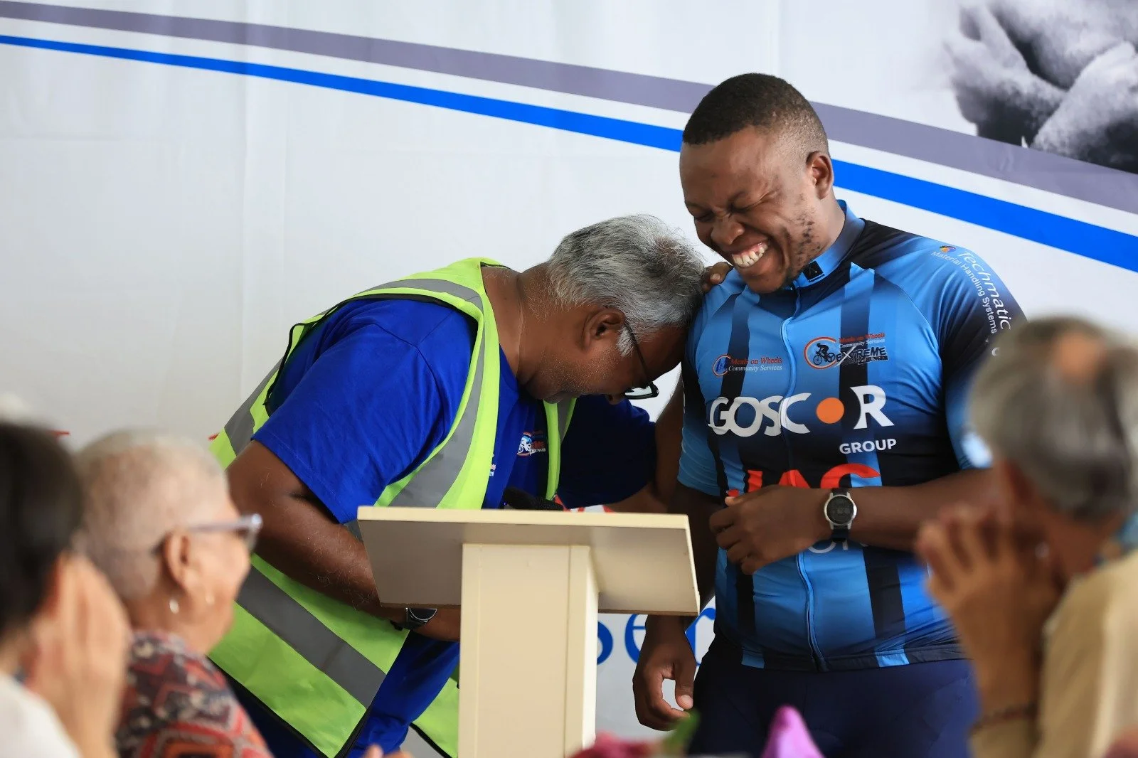 A man in a blue cycling jersey smiling as another man leans forward with his head on the cyclist's chest, both sharing a joyful moment at an indoor event surrounded by seated people.