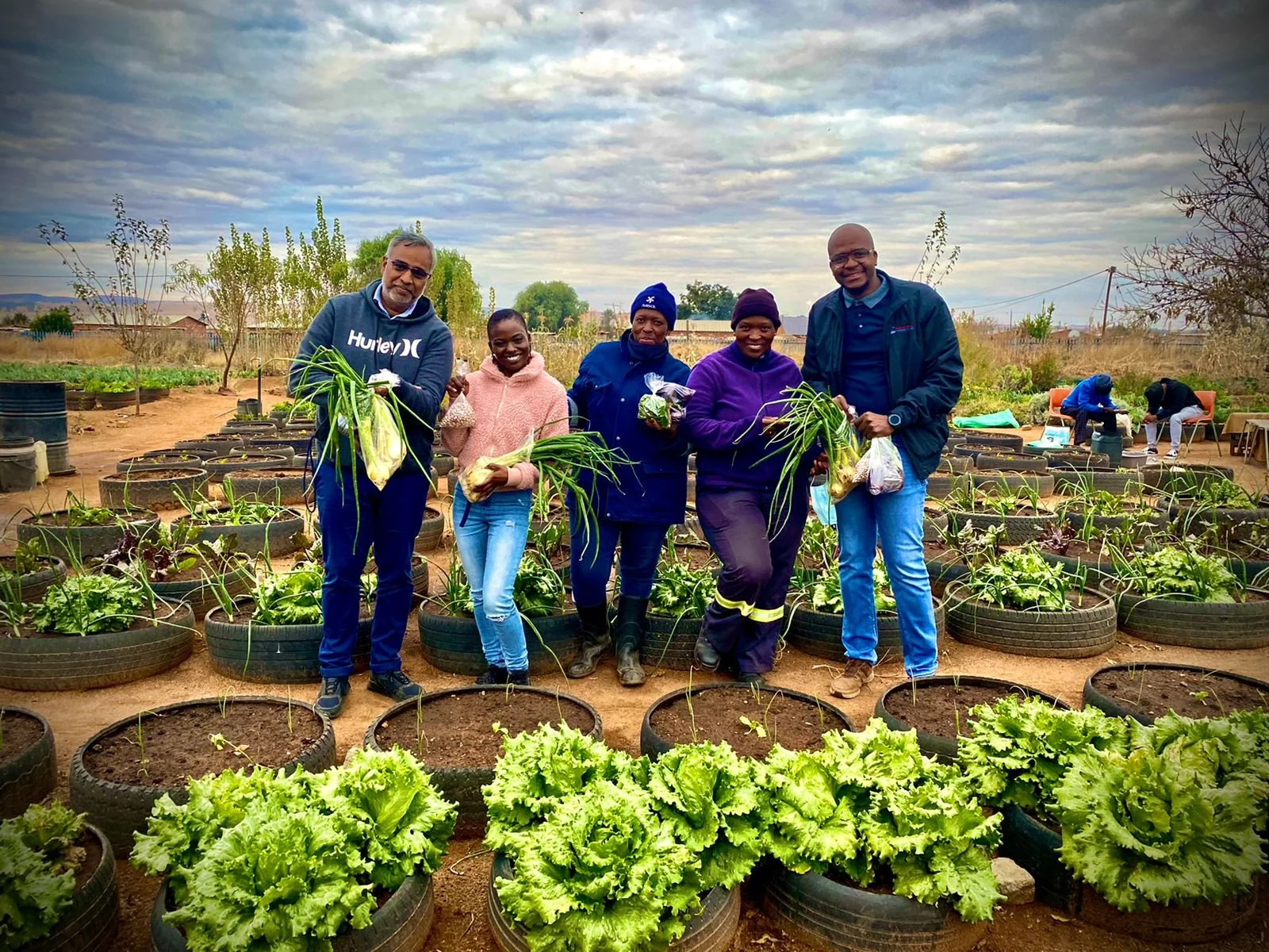 Group of five people standing in a garden with plants in tires, holding harvested vegetables, with cloudy sky and other gardeners in the background.