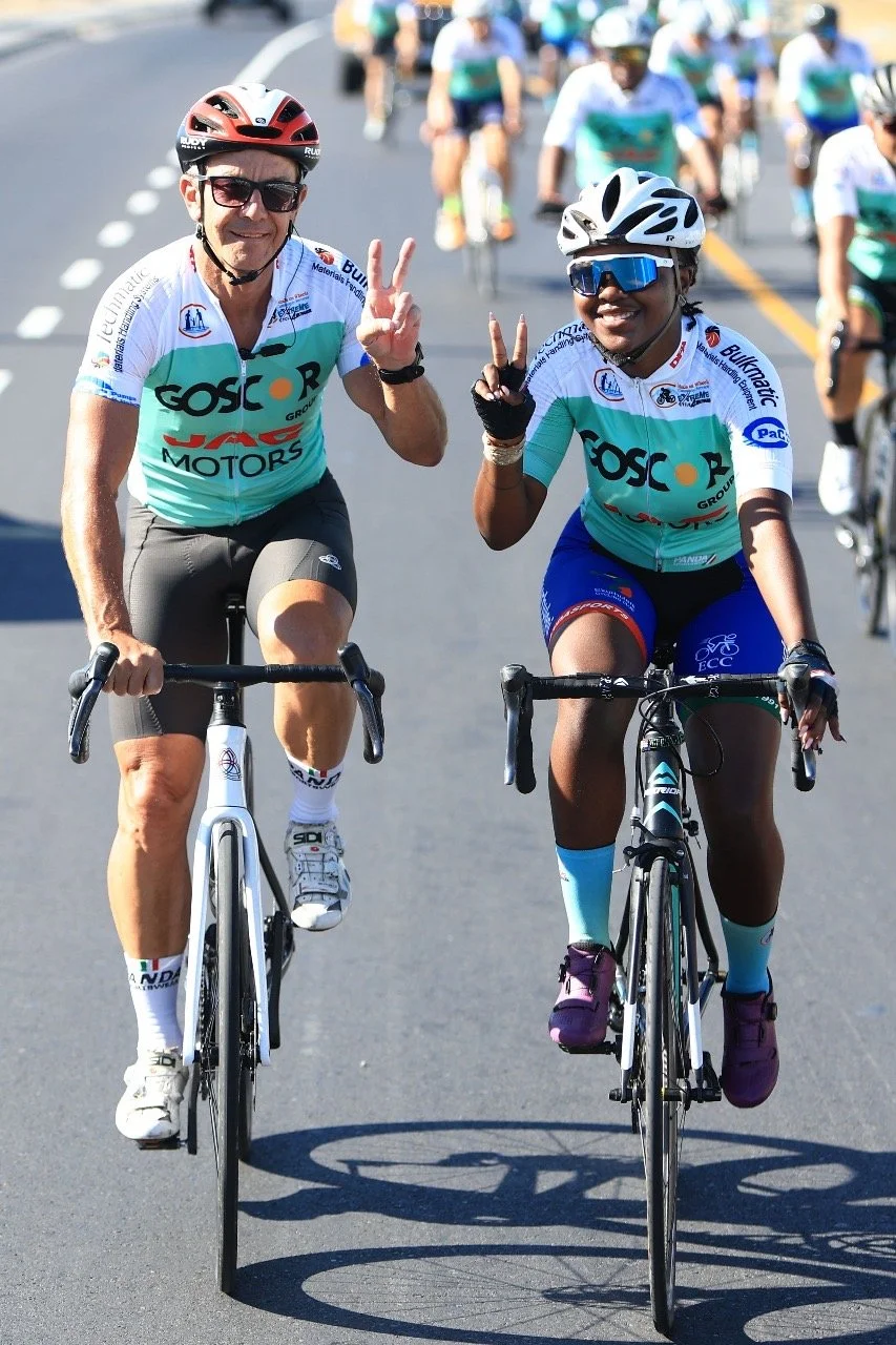 Two cyclists wearing helmets and sunglasses, riding bikes on a road, making victory signs with their hands, surrounded by other cyclists in a race.