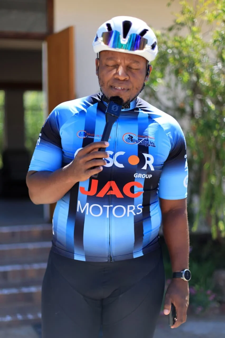 A man in a blue cycling jersey and black pants, wearing a white helmet, holding a microphone and a smartphone outdoors.