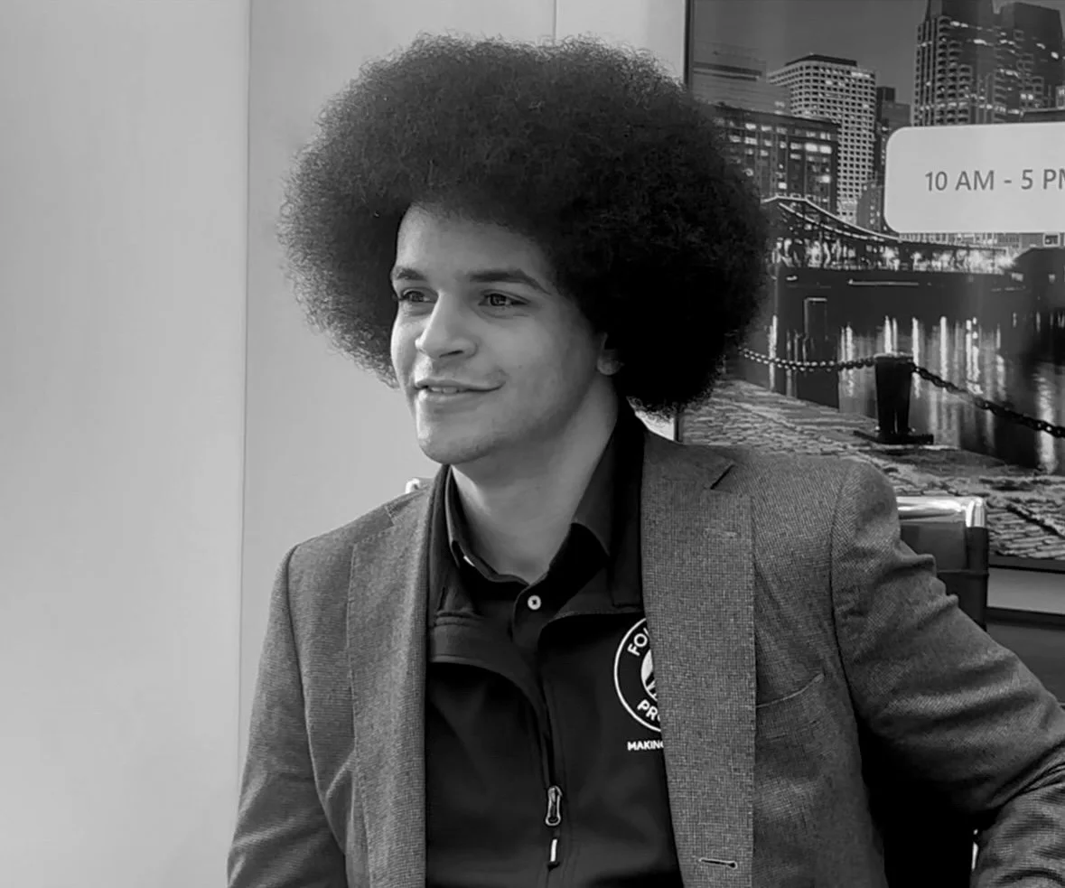 A young man with an afro hairstyle wearing a blazer and a jacket with a logo, sitting indoors in front of a cityscape mural.