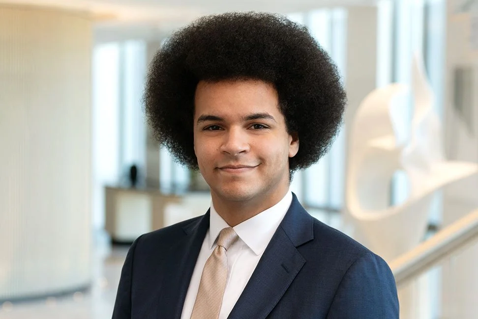Portrait of a young man with a large afro hairstyle, wearing a suit and tie, smiling in an office setting.