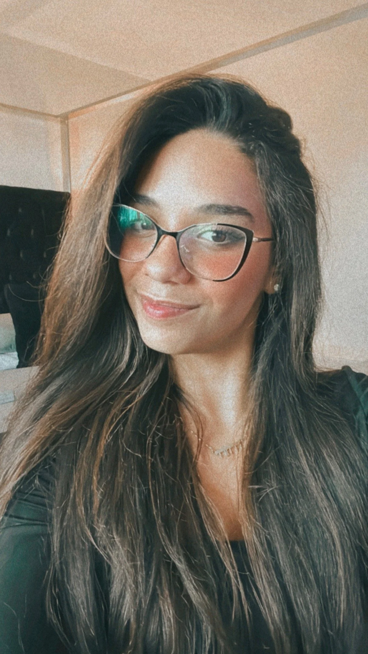 A woman with long, wavy brown hair, wearing glasses, a black top, and a gold necklace, smiling and taking a selfie indoors.