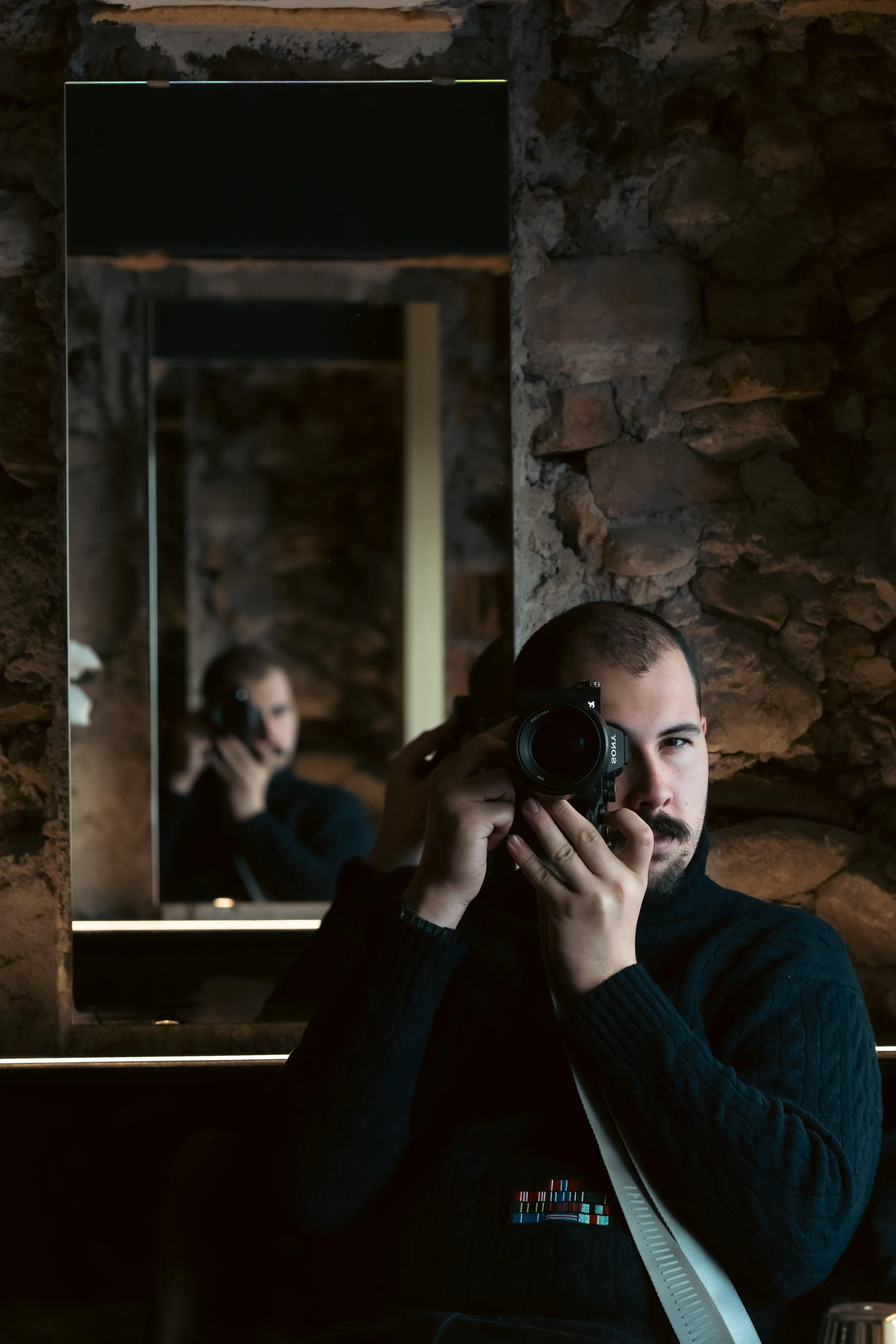 A man is taking a selfie in a mirror with a camera, with a woman in the background also holding a camera, in a room with exposed brick walls.