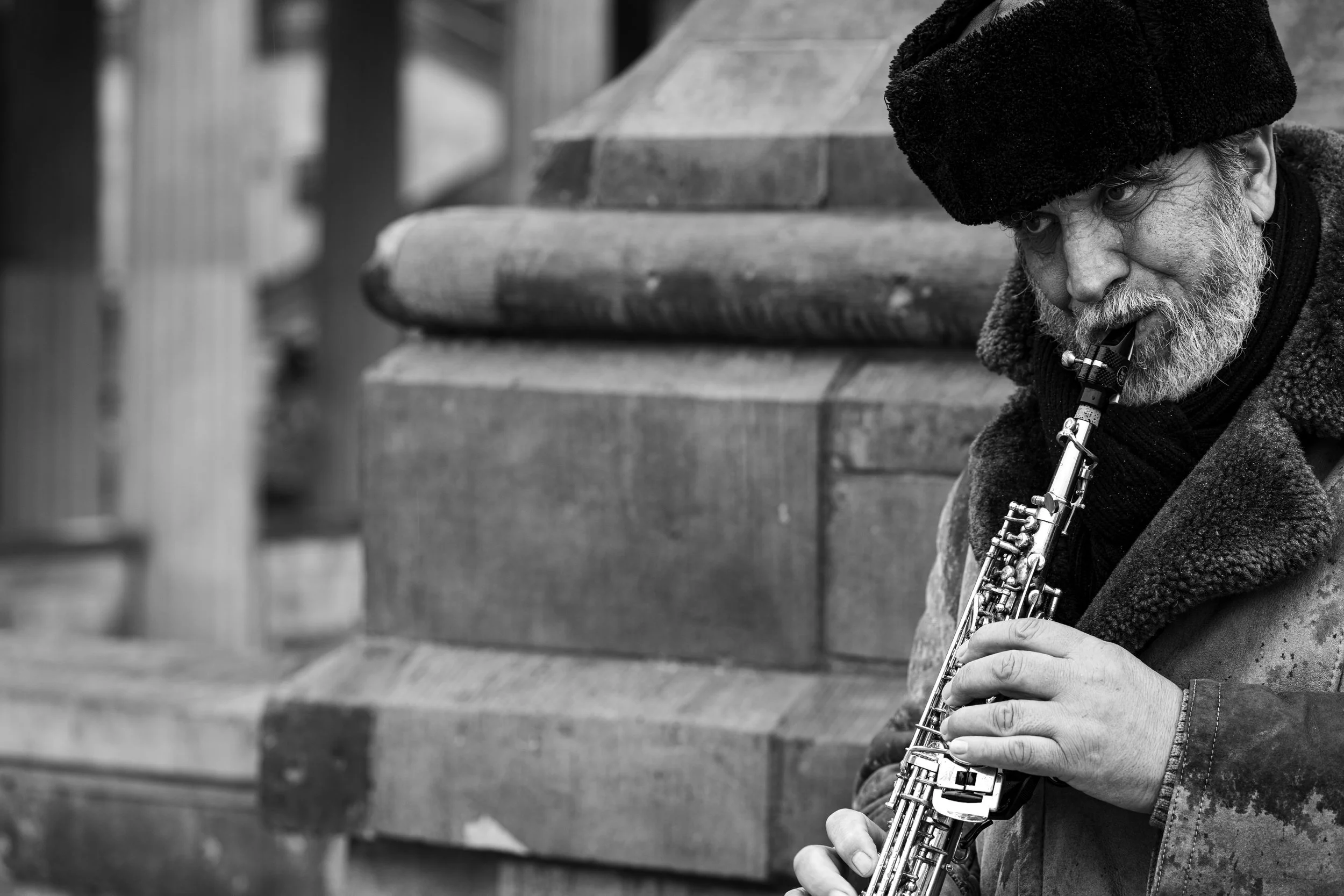 A black-and-white photo of an elderly man with a beard, wearing a fur hat, playing a saxophone outdoors near a wooden structure.