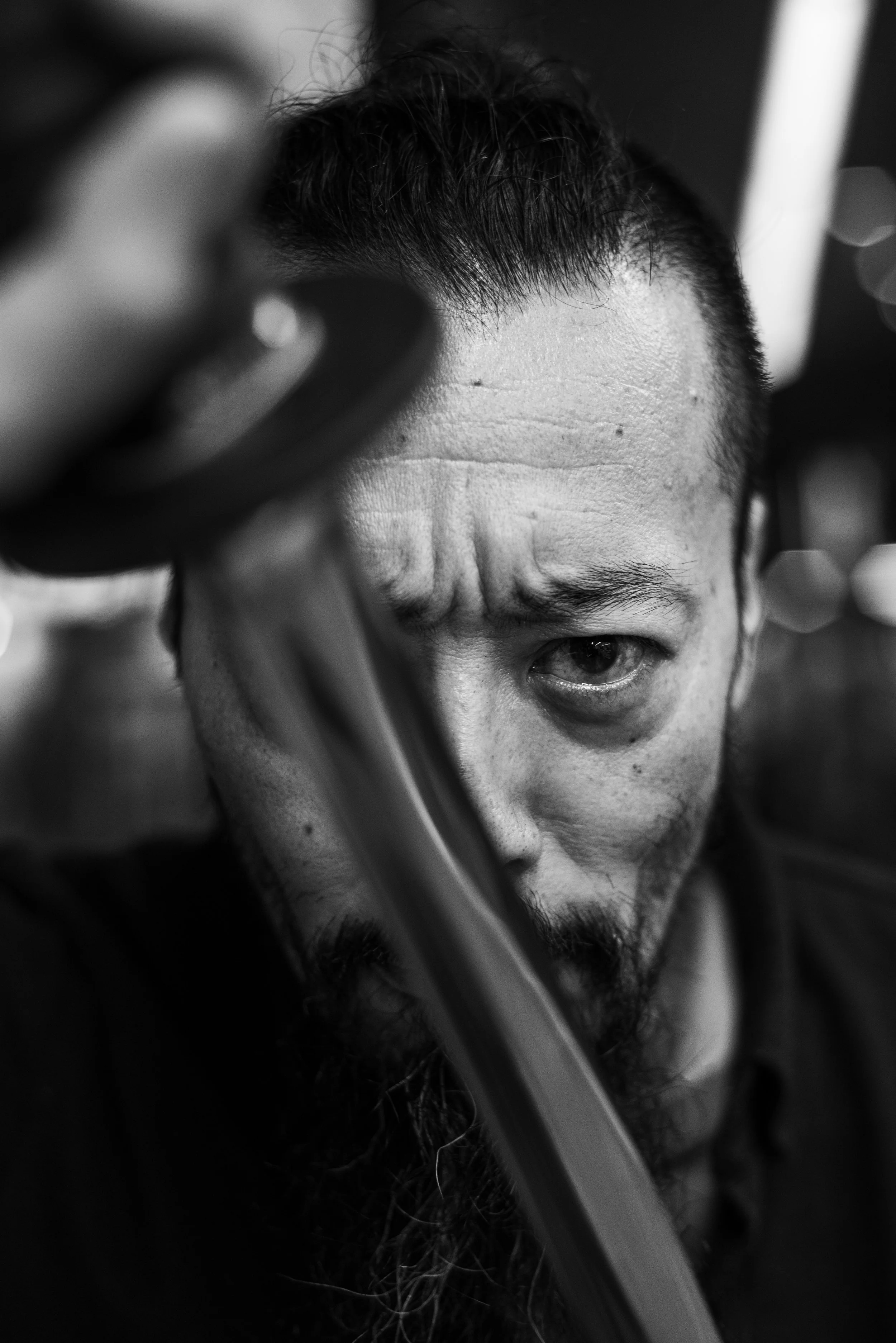 Close-up black and white photo of a man with short hair and a beard, looking directly at the camera with his forehead slightly furrowed, hand partially covering his face.