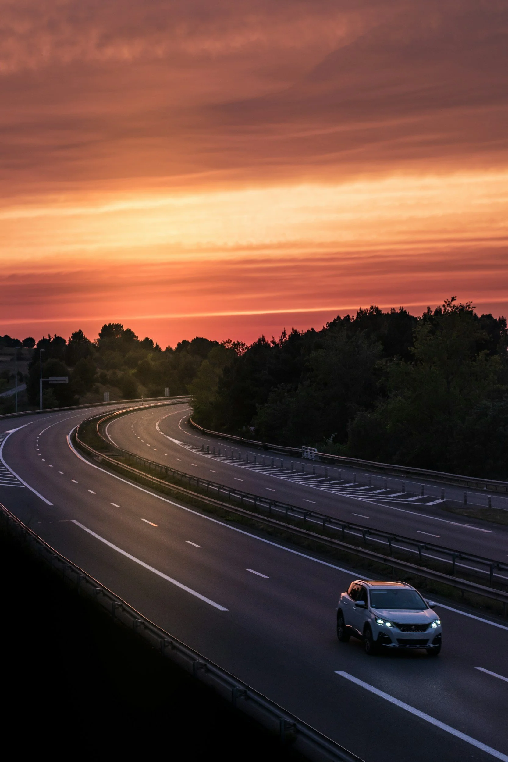 A white SUV driving on a curving highway at sunset with colorful orange and pink sky and forested area on the roadside.