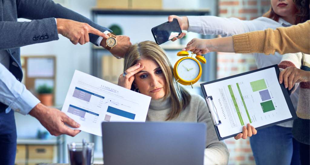 A woman looks stressed at her desk surrounded by multiple people pointing at her with documents, a phone, an alarm clock, and a laptop.
