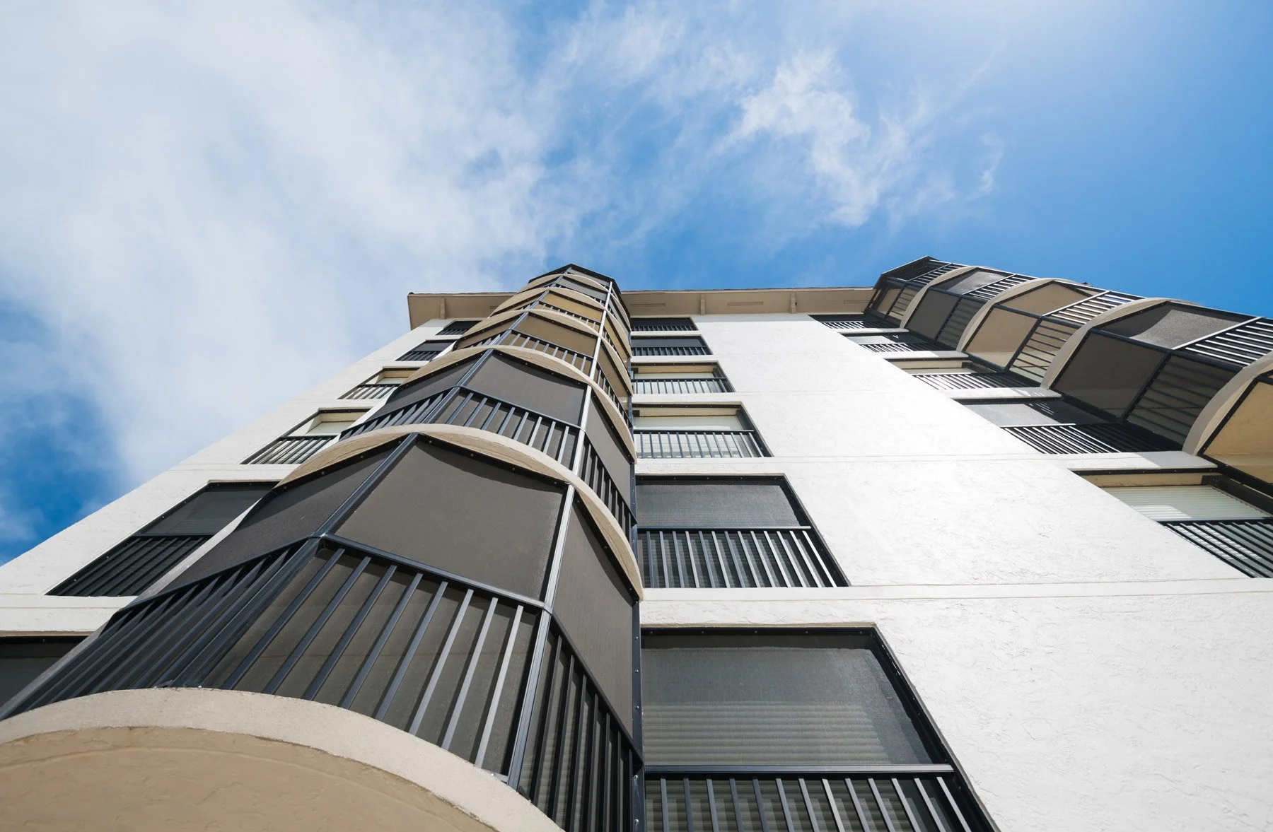 Looking up at a modern apartment building with multiple floors, black balconies, and large windows, against a partly cloudy blue sky.