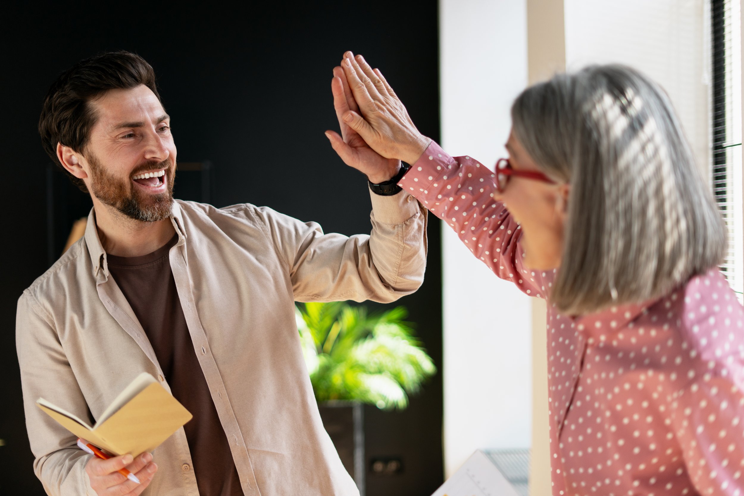 Man and woman giving each other a high five indoors, woman with gray hair and glasses, man with dark hair and beard, man holding a book and a pen.