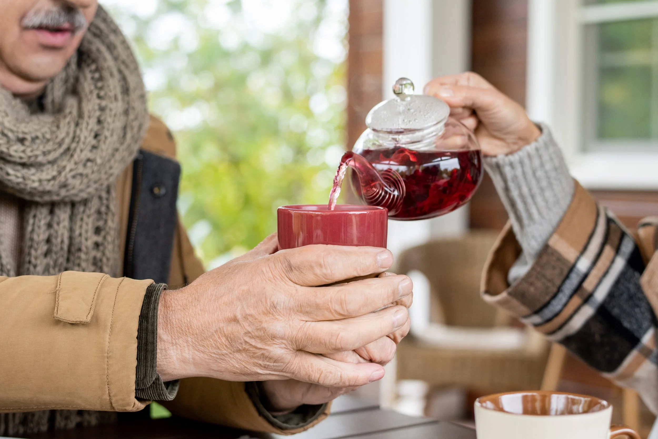 Person pouring red tea from a glass teapot into a red mug, with another person holding the mug, in a cozy, well-lit room with a green leafy view outside.