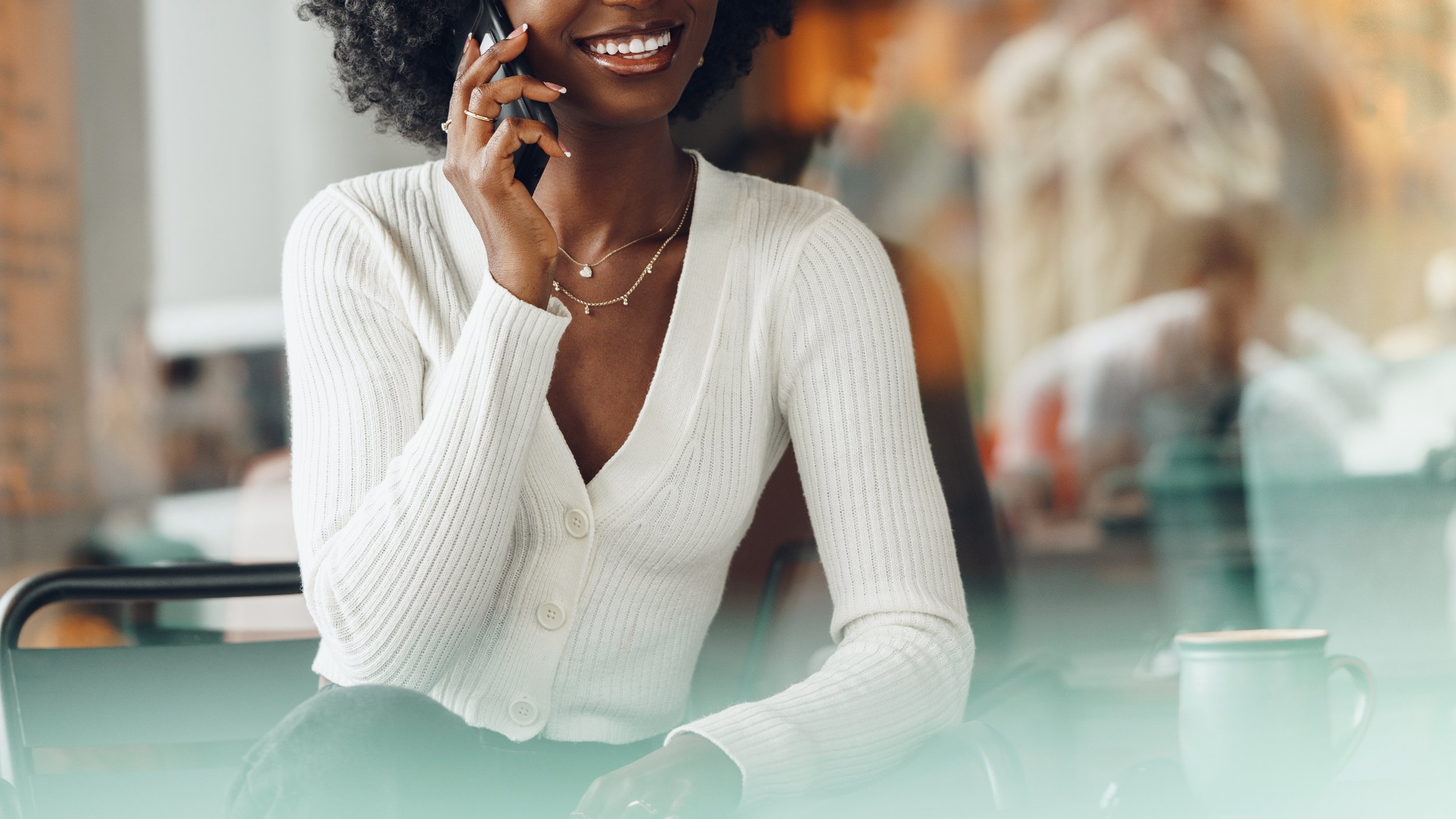 A woman with dark skin, curly hair, and a bright smile talking on a cellphone in a casual setting, wearing a white buttoned sweater and layered necklaces.