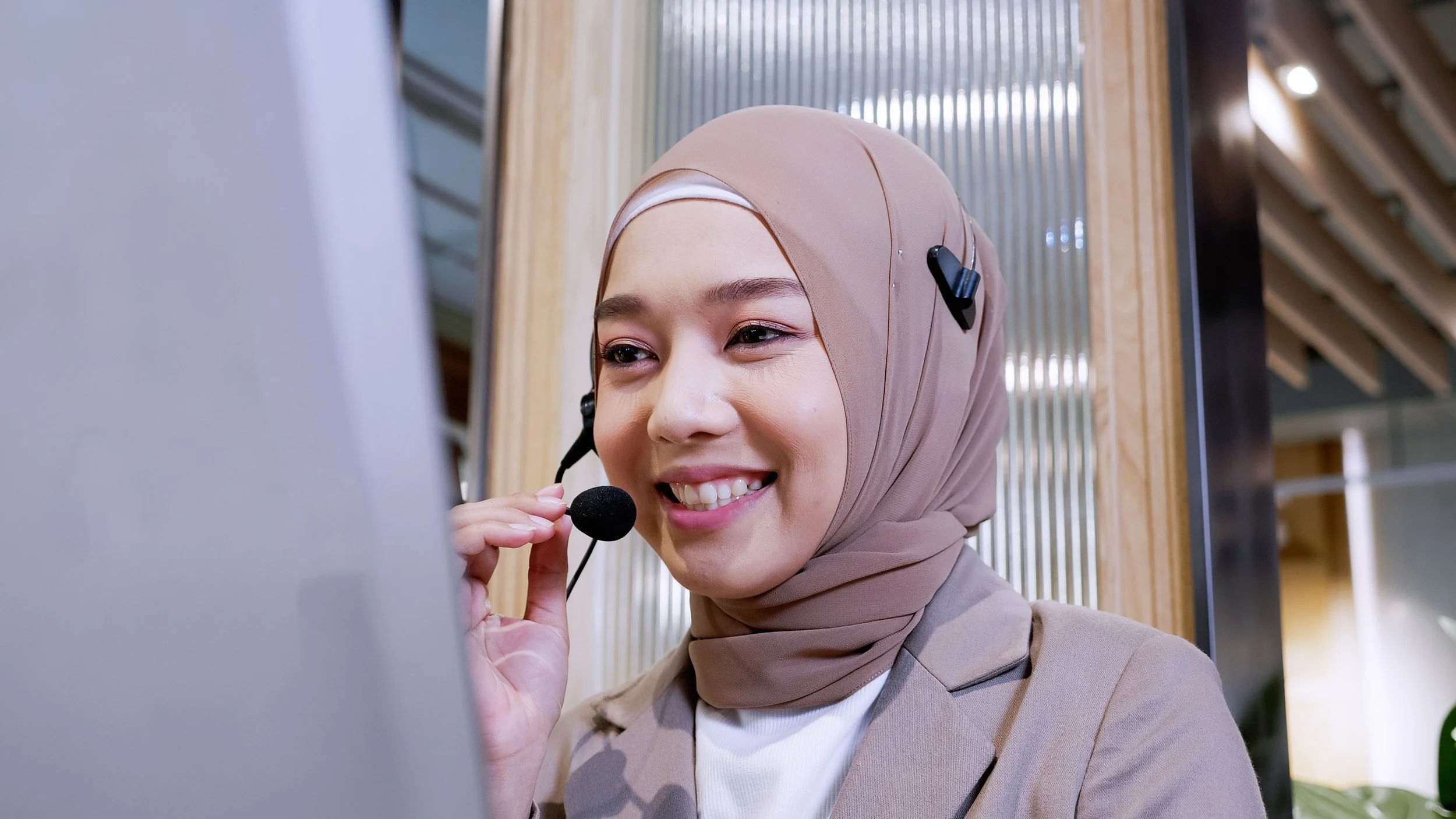 A woman wearing a beige hijab and a beige blazer smiling while adjusting a headset with a microphone at her ear, in a modern indoor setting.