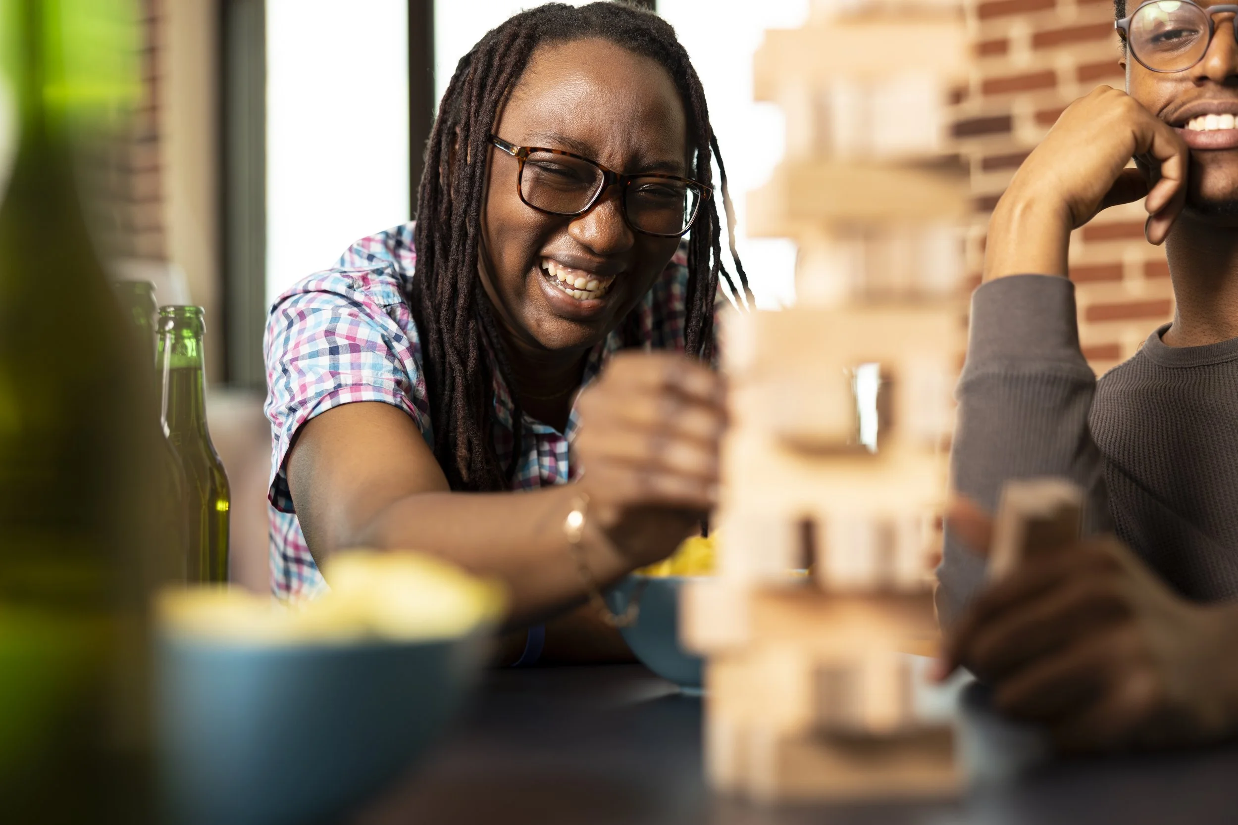 Two friends laughing and enjoying each other's company in a cozy indoor setting with brick walls. One person with dreadlocks and glasses is smiling broadly, while the other person, partially visible, is resting their chin on their hand, also smiling.