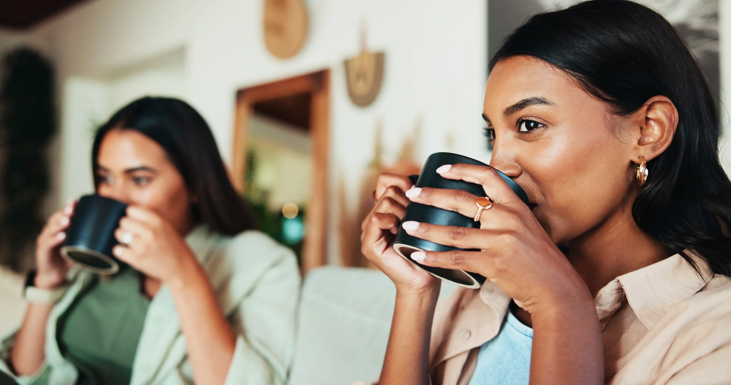 Two women enjoying hot drinks together indoors, one with dark hair and gold earrings, the other with dark hair and a ring, both smiling.