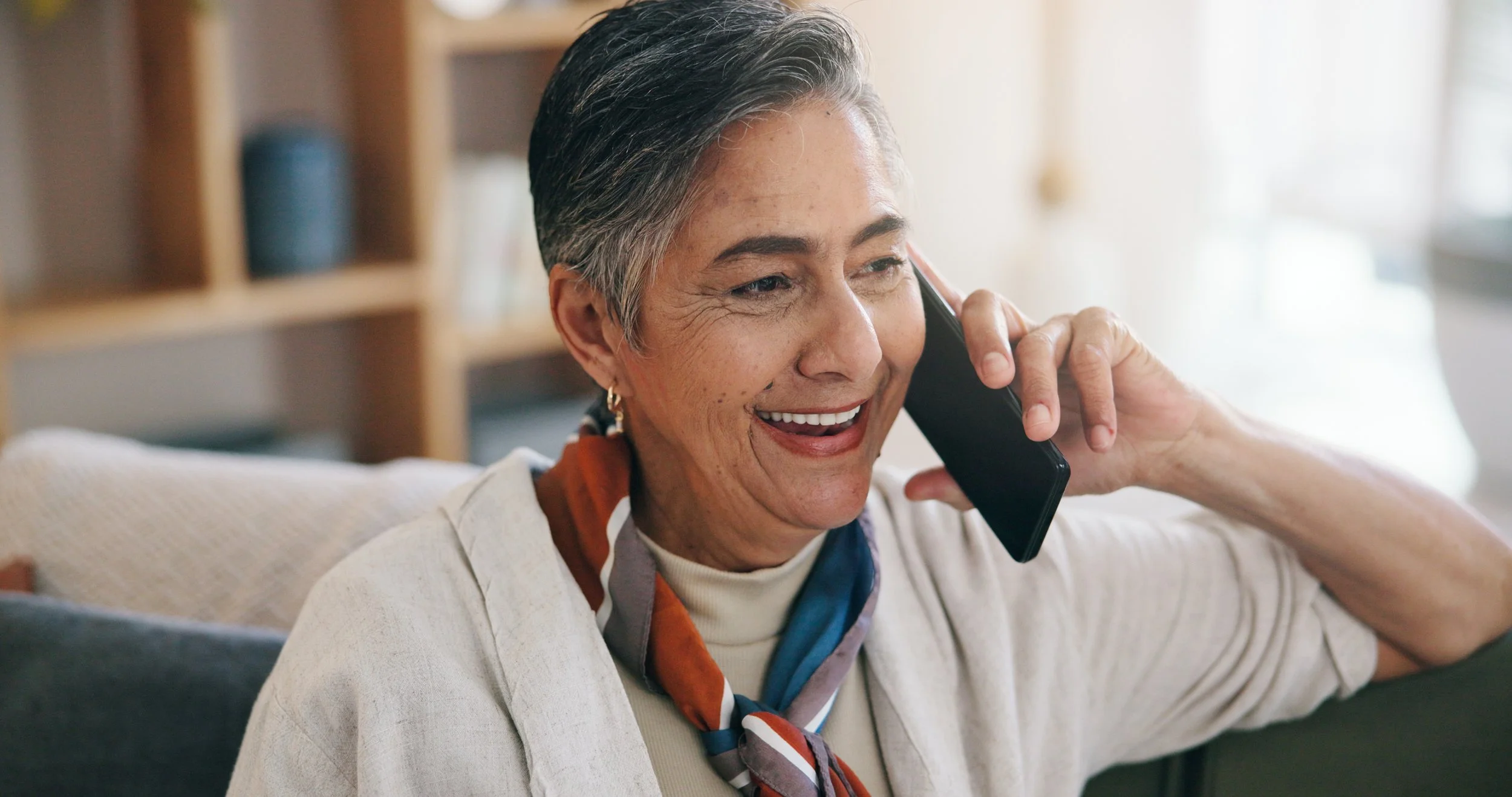 A smiling woman with short gray hair talking on a cell phone, sitting on a couch in a cozy room with a bookshelf in the background.