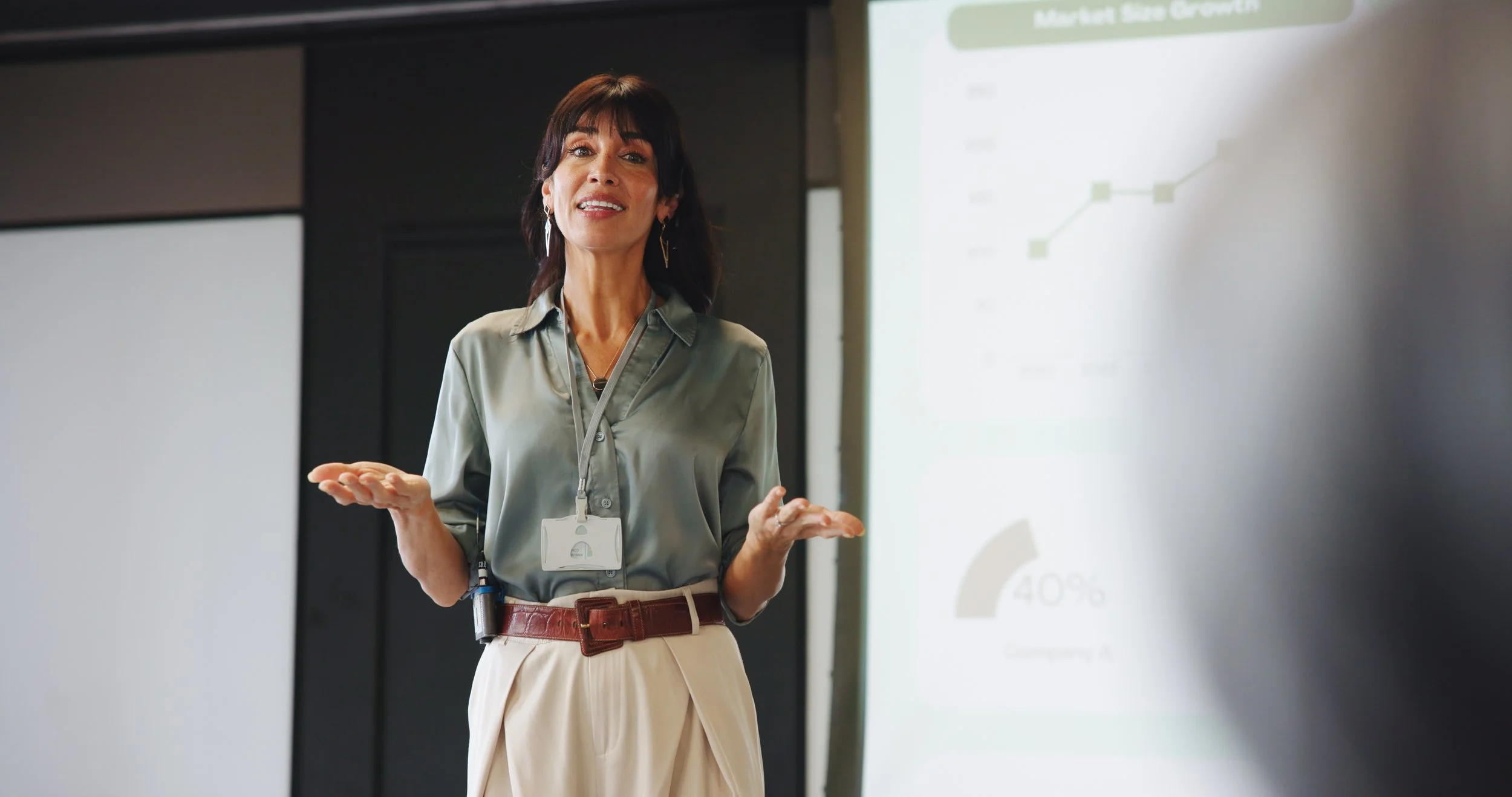 A woman in business attire giving a presentation in a conference room with a projection screen displaying a chart.