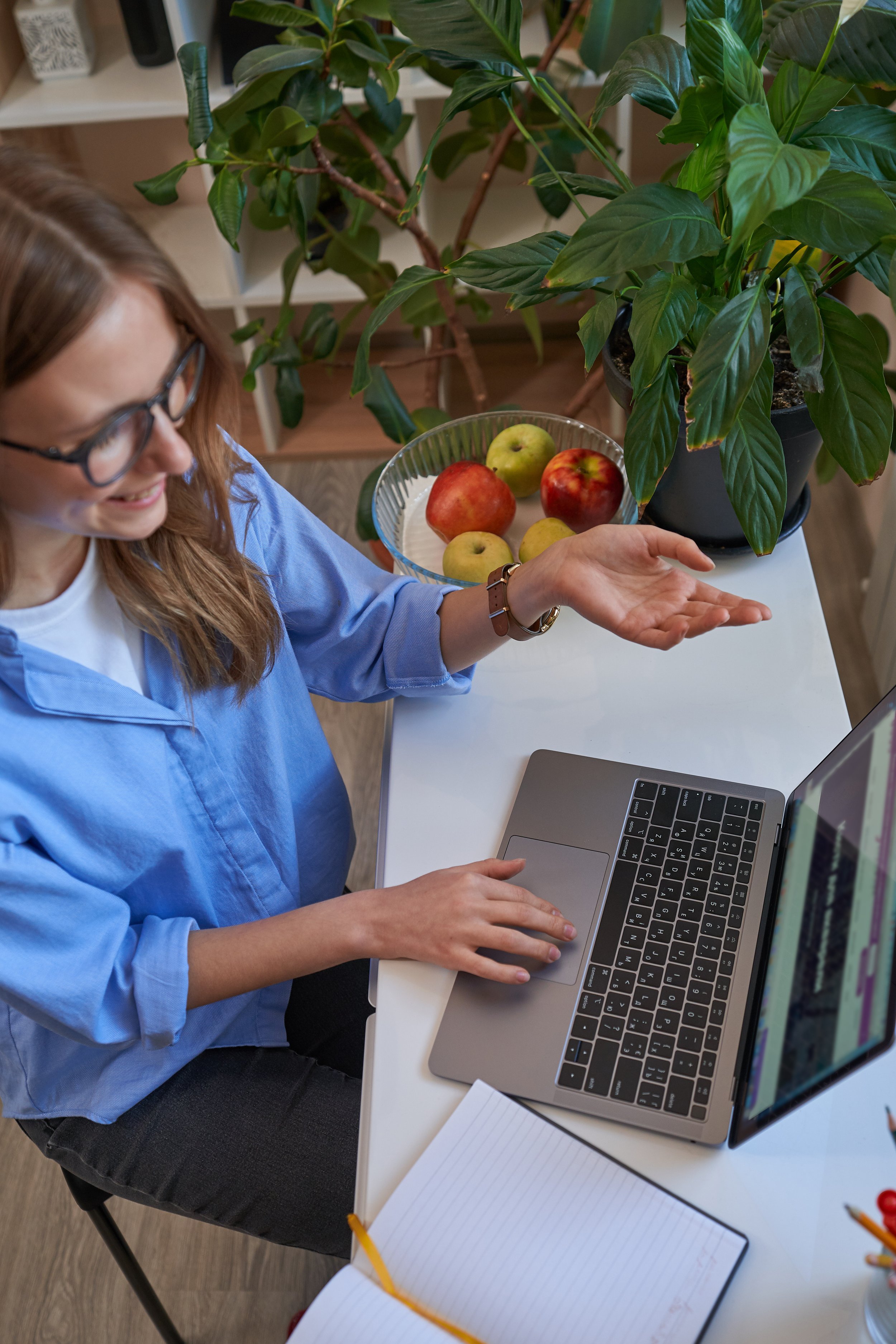 A woman with glasses, wearing a light blue shirt, sits at a white desk using a laptop. On the desk, there is a notepad with a yellow pencil, a notebook, a water bottle, a bowl of fruit with apples, and a large green houseplant in a black pot.