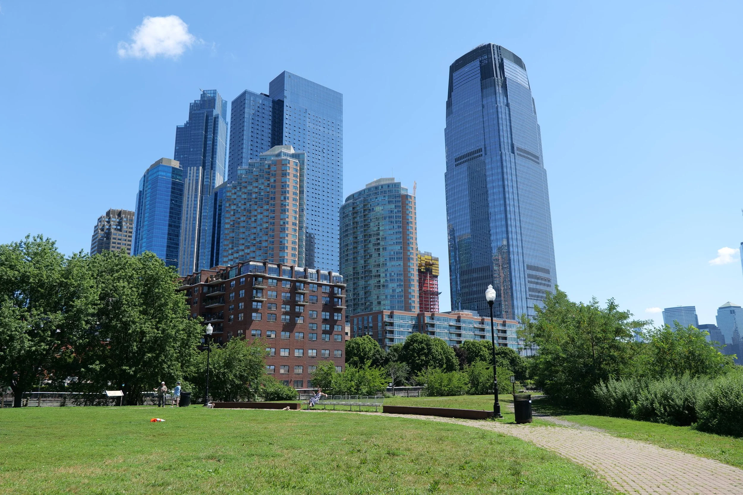 City skyline with tall modern skyscrapers viewed from a park with green trees, people, benches, street lamps, and a clear blue sky.