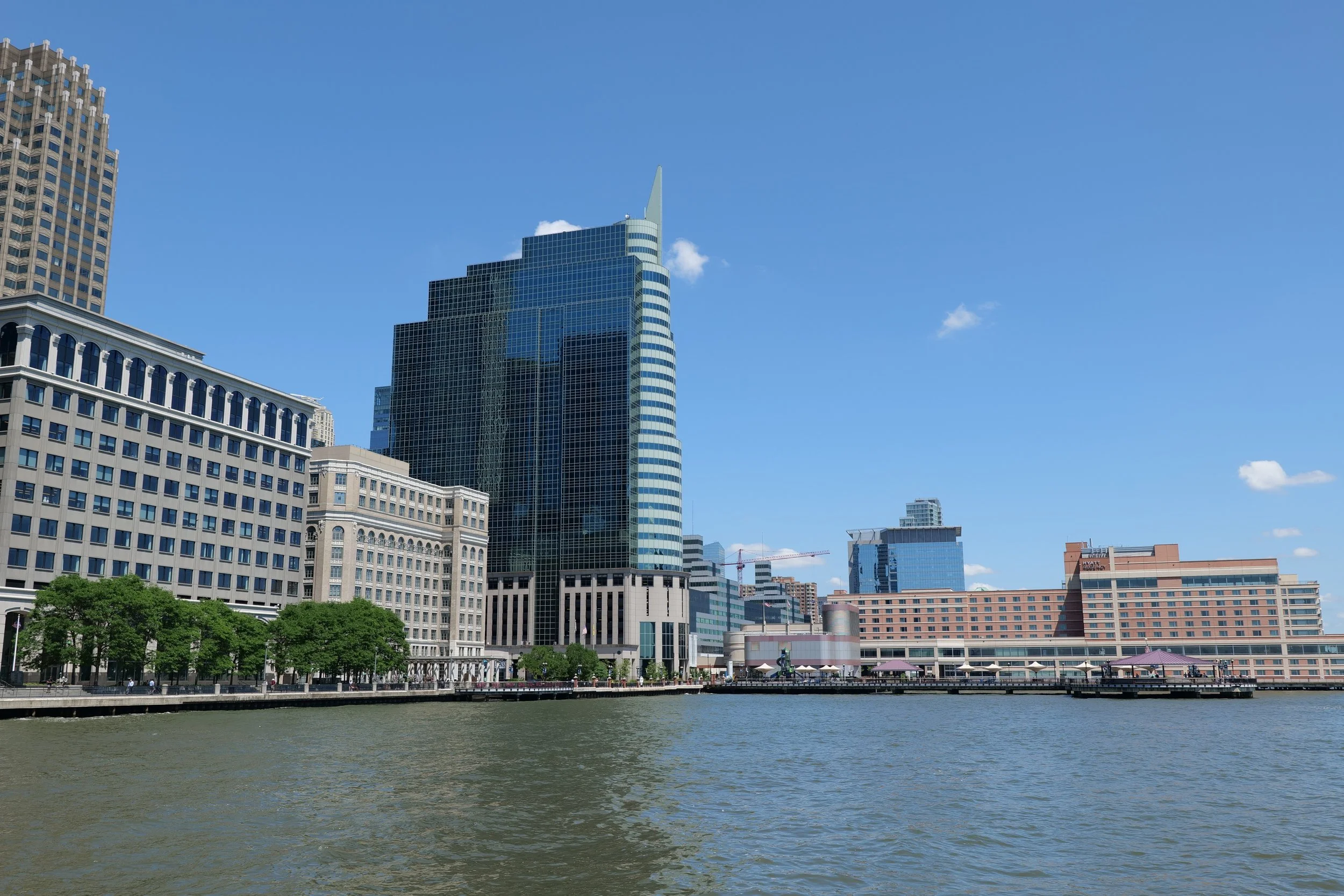City skyline with modern glass skyscraper, older buildings, trees, and body of water in foreground under a clear blue sky.