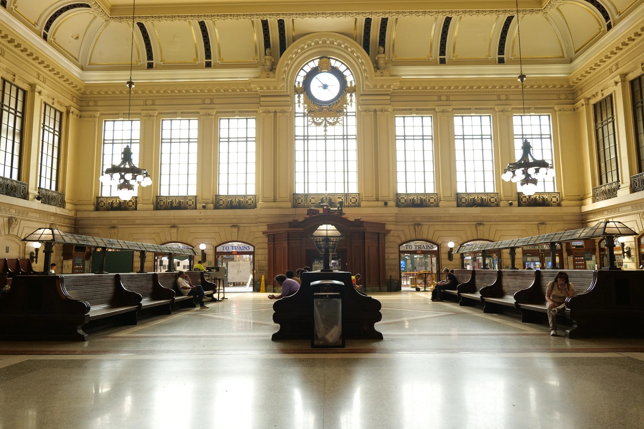 Interior view of a historic train station with high arched ceilings, large windows, a clock in the center, and seating areas with people waiting.