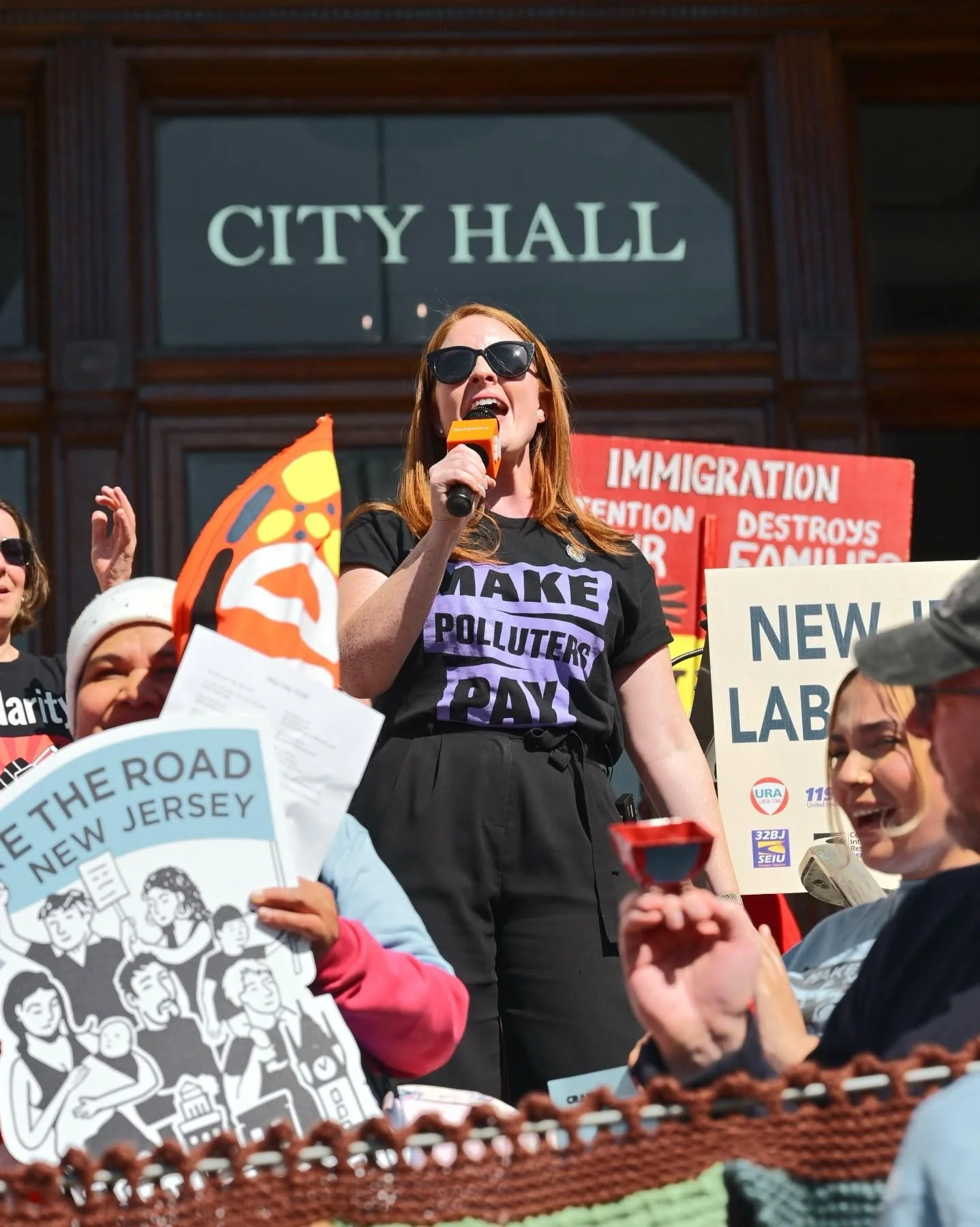 Assemblywoman Katie Brennan speaking into a microphone during a protest outside a city hall building, holding a sign that reads 'MAKE POLLUTERS PAY'. Several other protesters are visible holding signs and pamphlets.