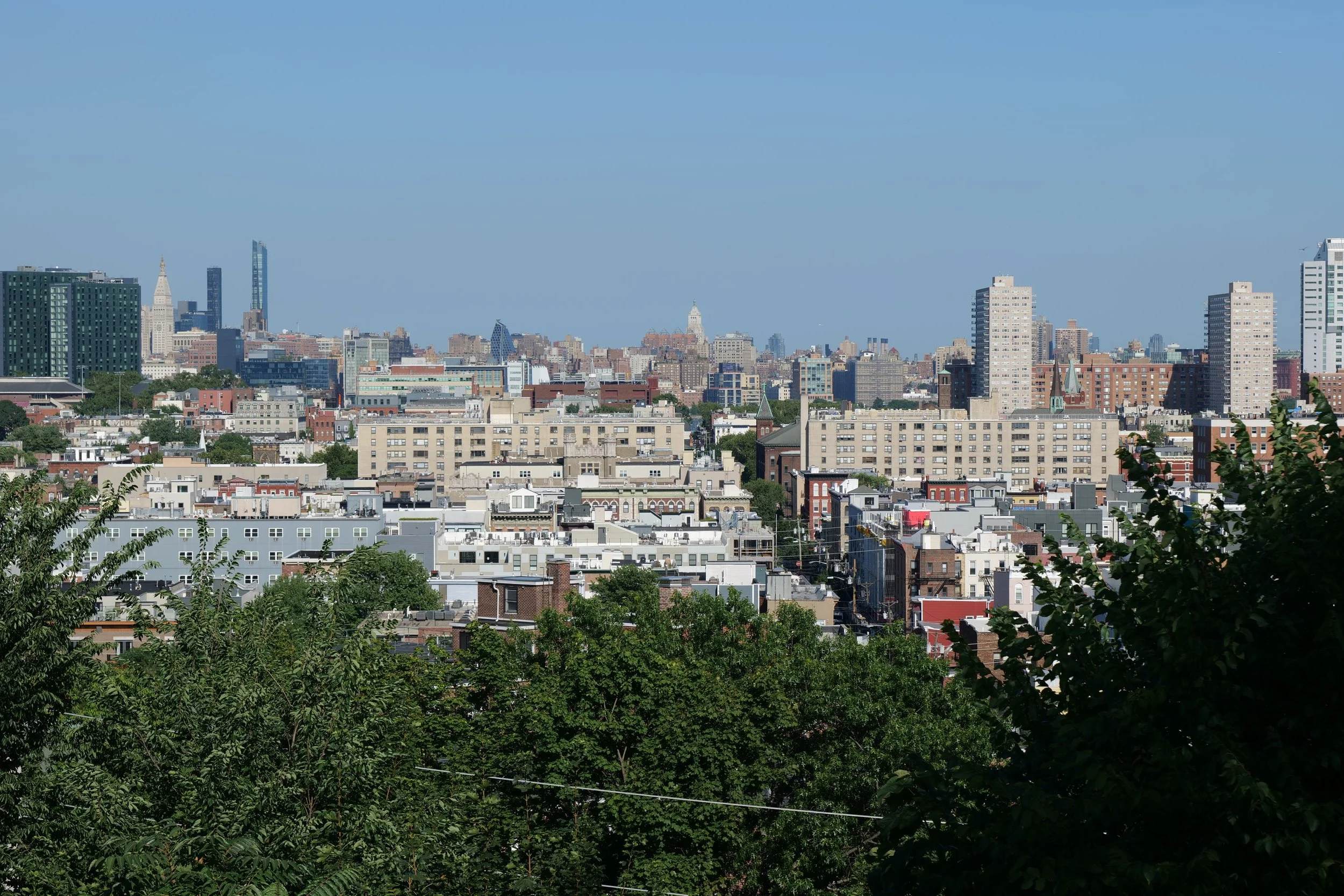 Skyline of a city with tall buildings, some modern and some historic, with greenery in the foreground and a clear blue sky.