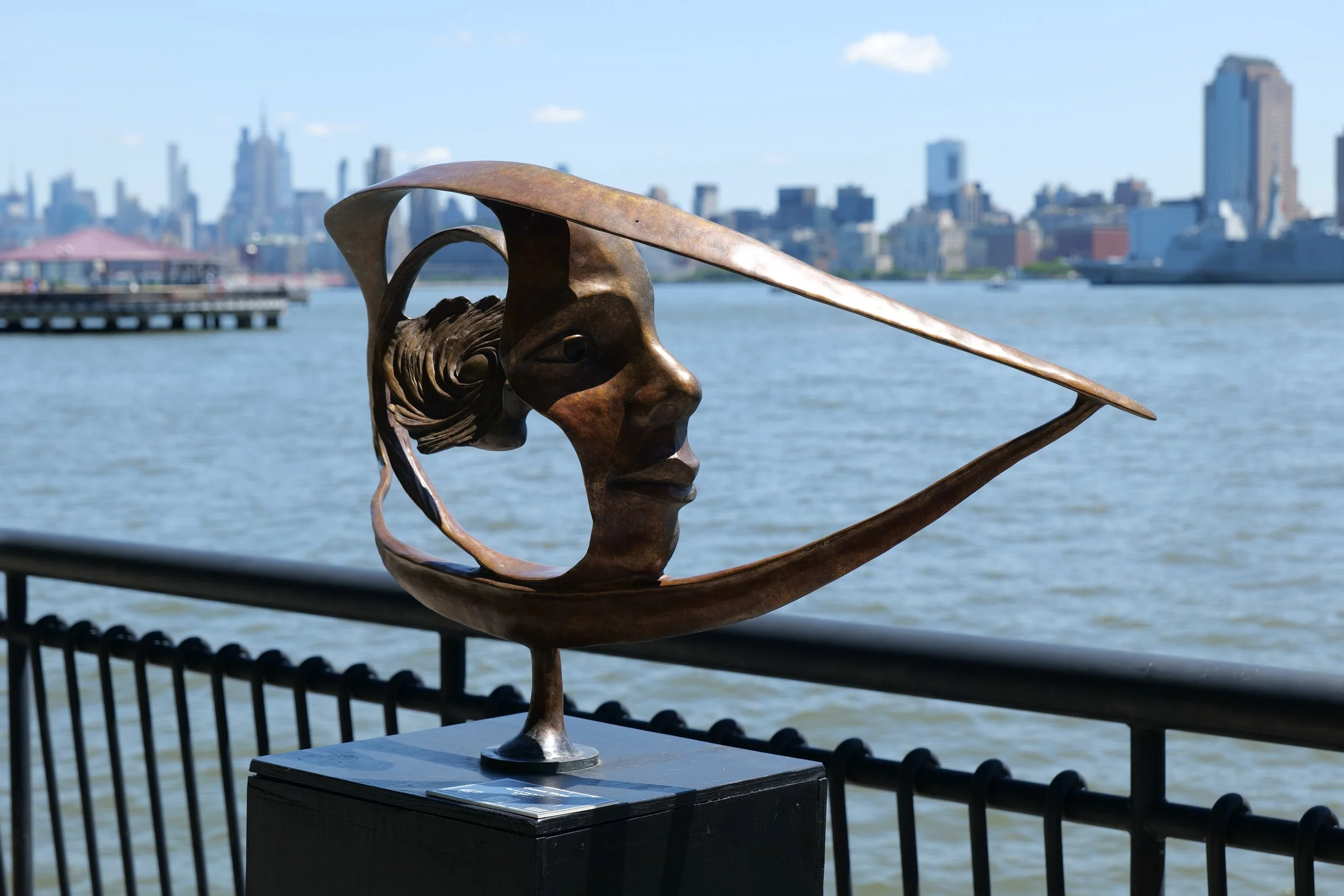 A metal sculpture of a human face with a long, flat extension resembling a sword or blade on the forehead, set against a backdrop of a city skyline and river, with part of a black railing and pedestal in the foreground.