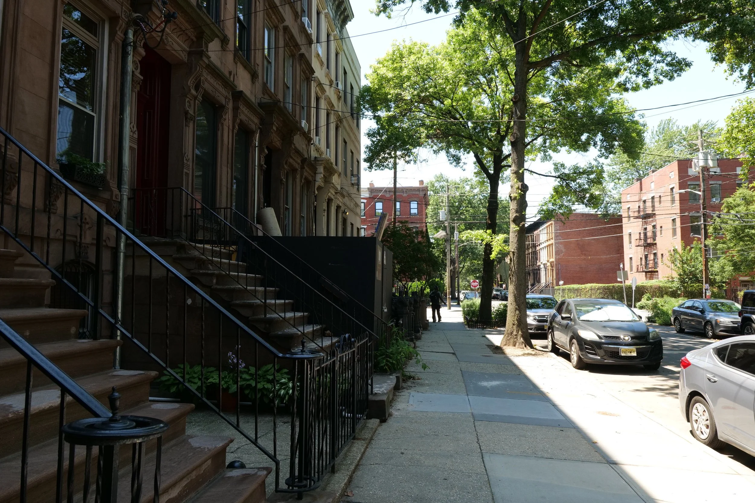 Residential street scene with brownstone row houses, trees, parked cars, and a person walking on the sidewalk during daytime.