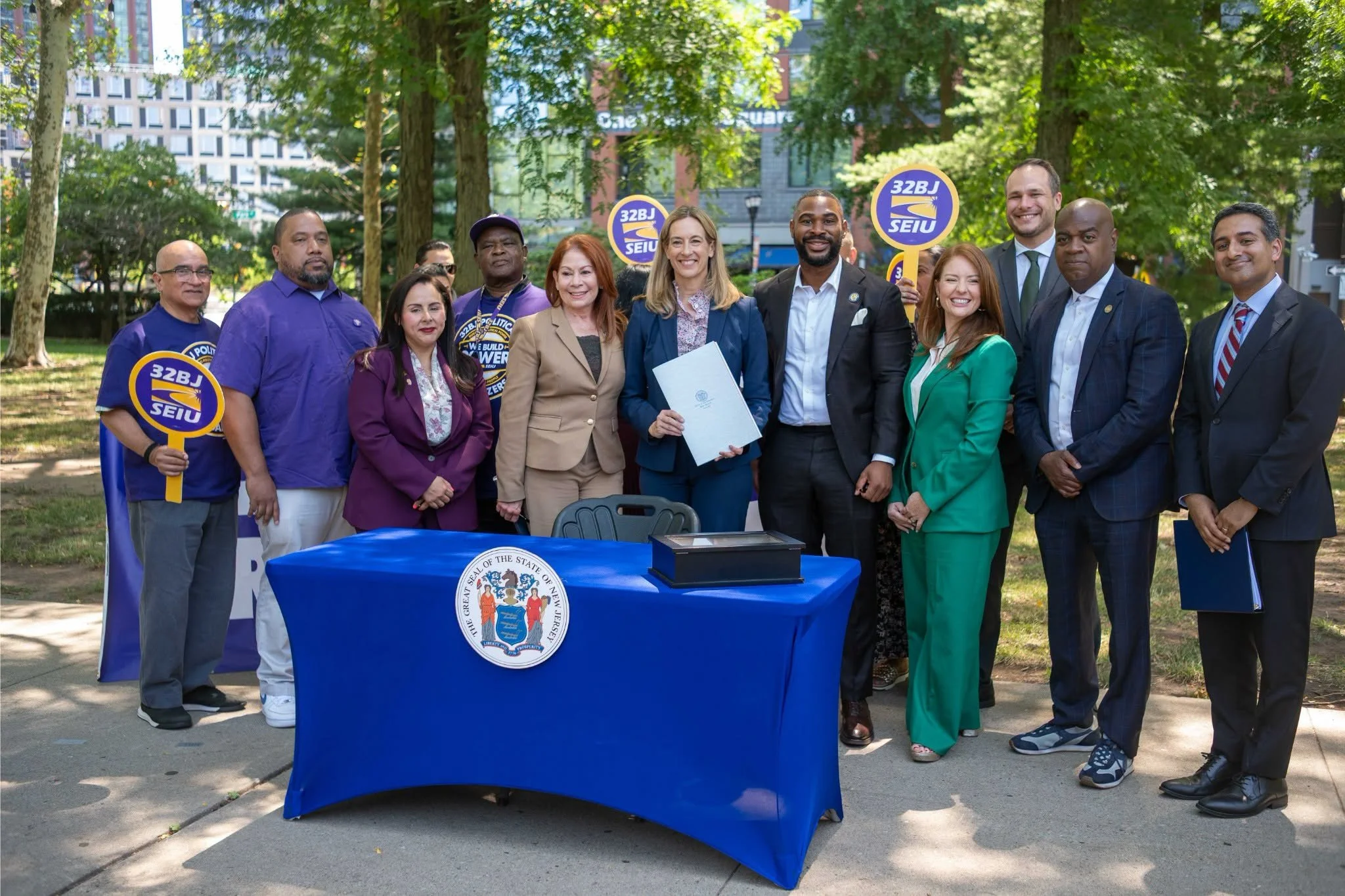 A group of diverse people standing together outdoors in a park, smiling and holding signs with the SEIU logo. There is a table with a blue cloth and the New Jersey seal.