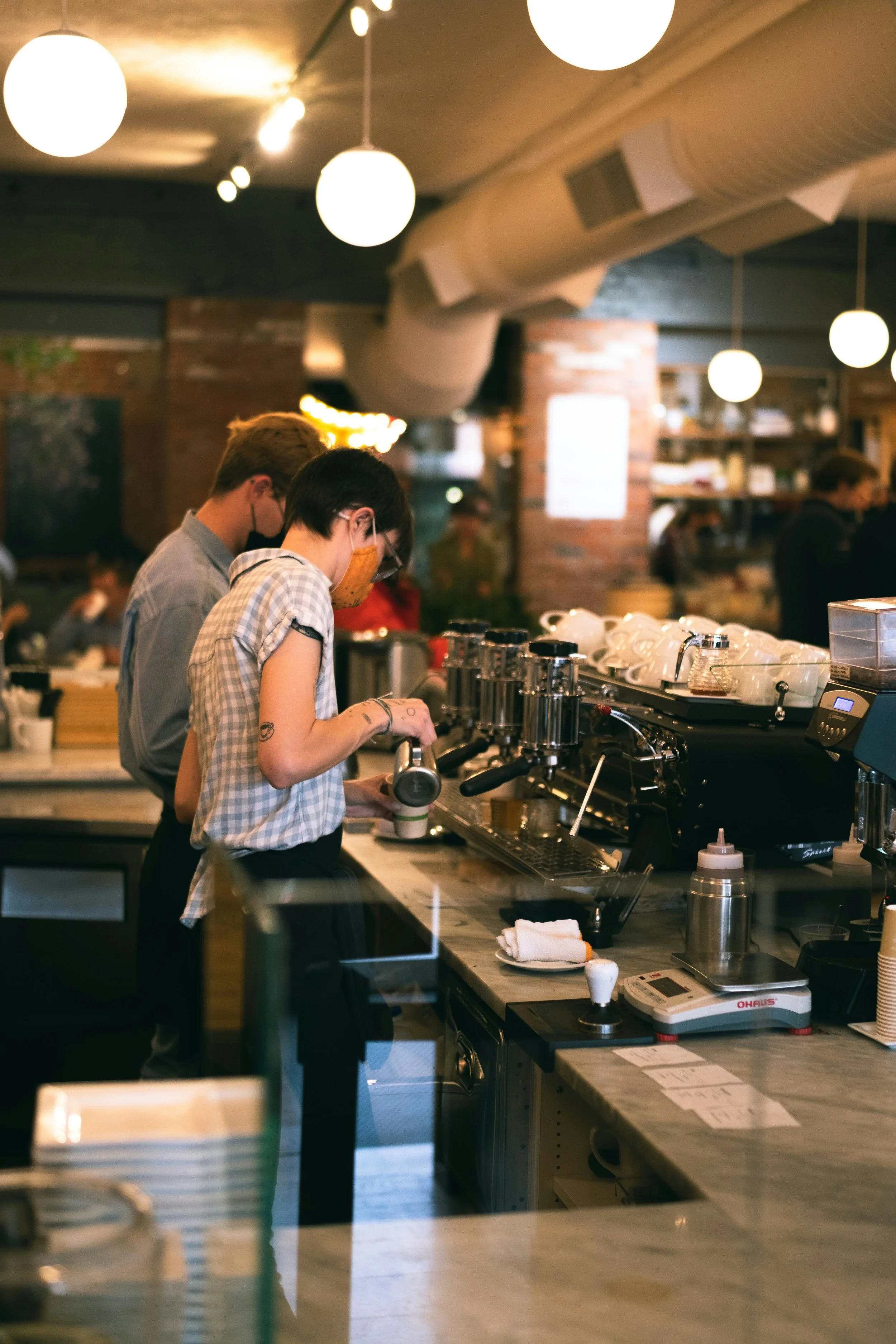 Two baristas wearing face masks working behind the counter at a coffee shop, with various coffee equipment and supplies on the counter and warm lighting overhead.