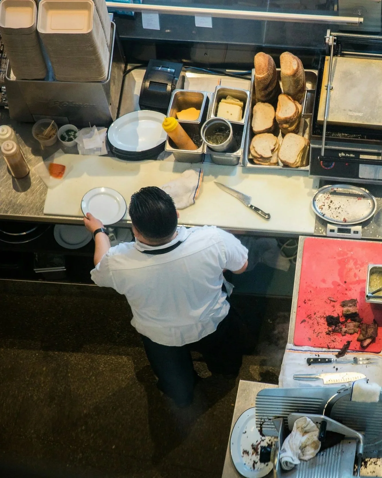 A chef standing at a kitchen counter with slices of bread, cheese, condiments, and a cutting board. There are bread slices stacked on a tray, and a pink cutting board with some cooked meat. The chef is holding an empty plate, and there are various kitchen tools and containers around.