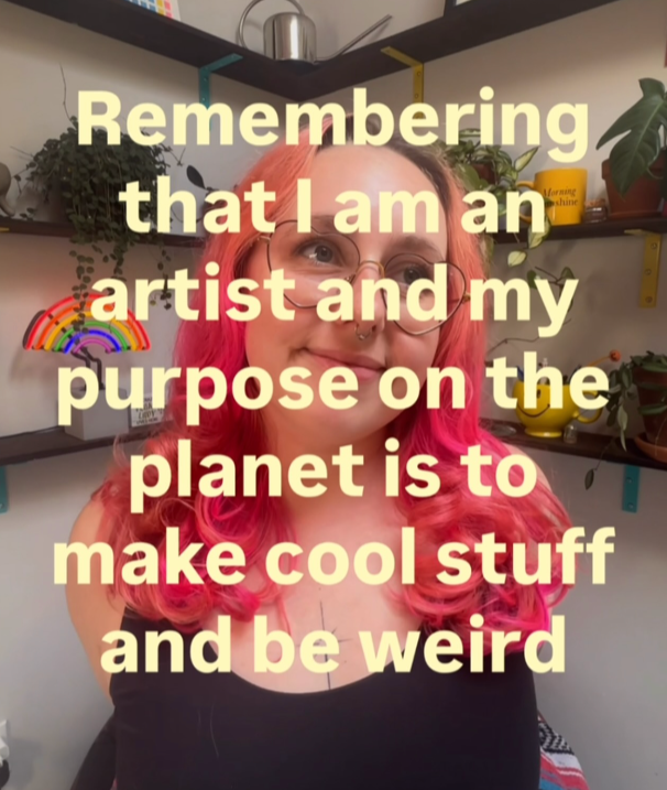 A woman with pink hair and glasses smiling in front of shelves with plants and decorative objects, overlaid with yellow text that reads: 'Remembering that I am an artist and my purpose on the planet is to make cool stuff and be weird.'