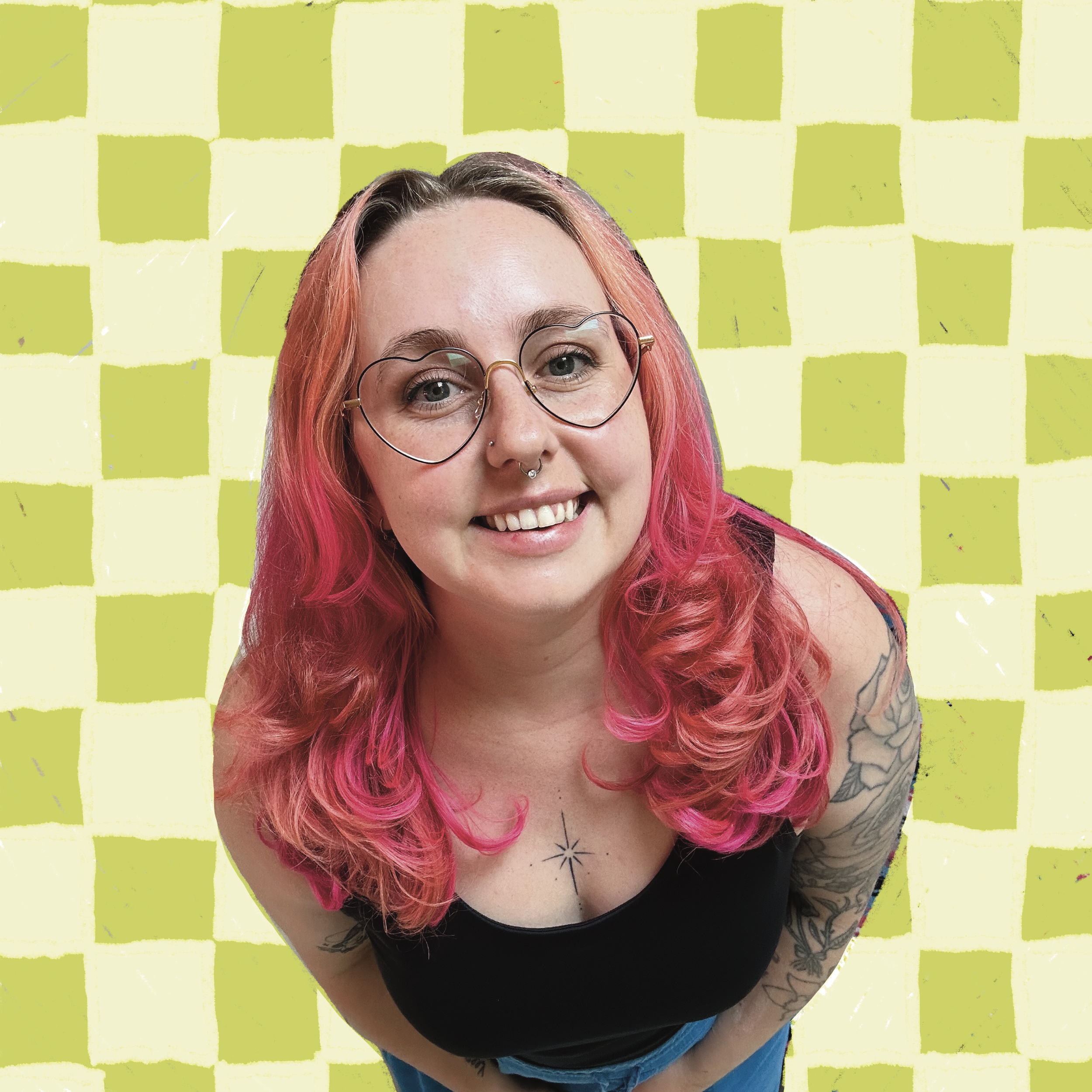 A woman with pink hair wearing glasses, a nose ring, and a black top, smiling at the camera against a green checkered background.