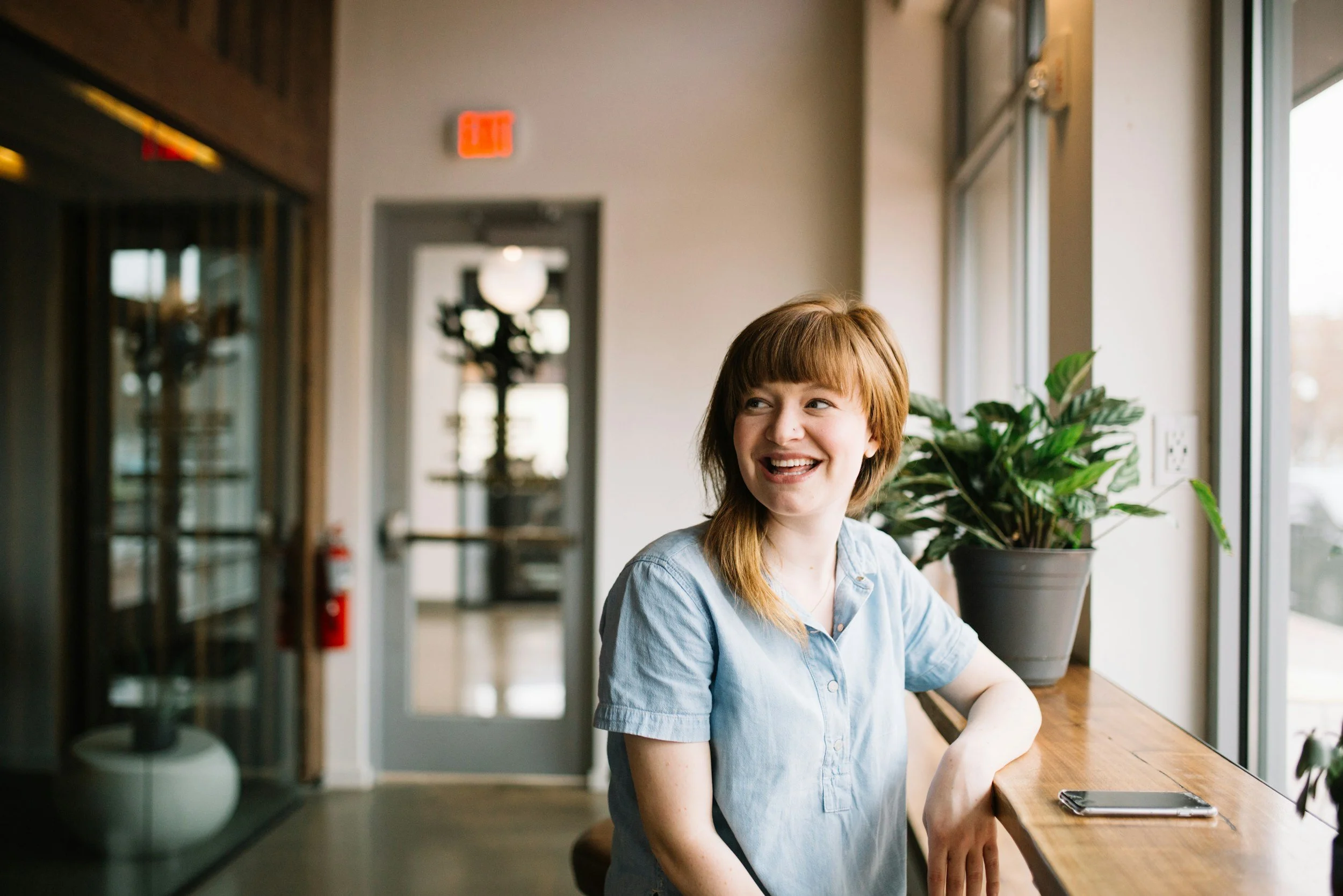 A woman with red hair and a blue shirt sitting at a wooden counter by a large window, smiling and looking to her right, with a potted plant beside her.