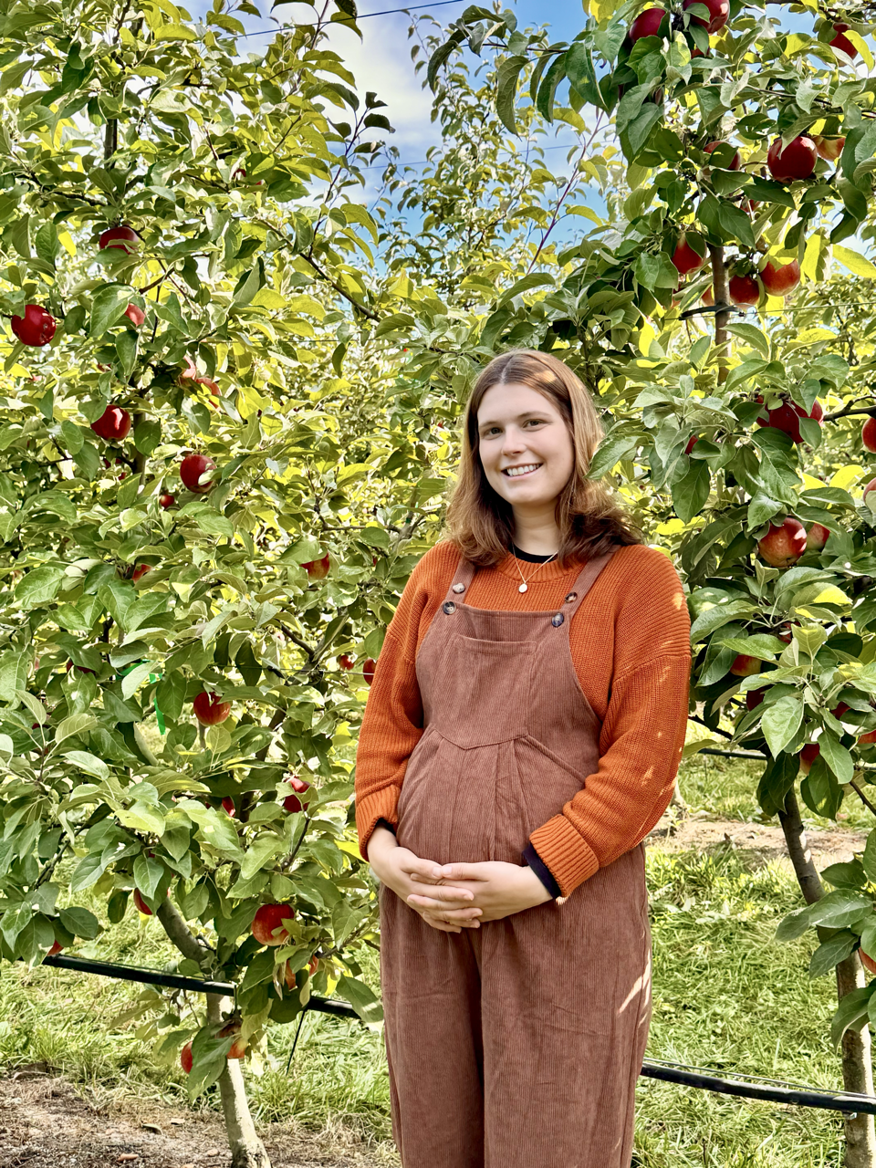 A young woman with brown hair smiling at the camera standing in an apple orchard with ripe red apples on trees and green leaves.