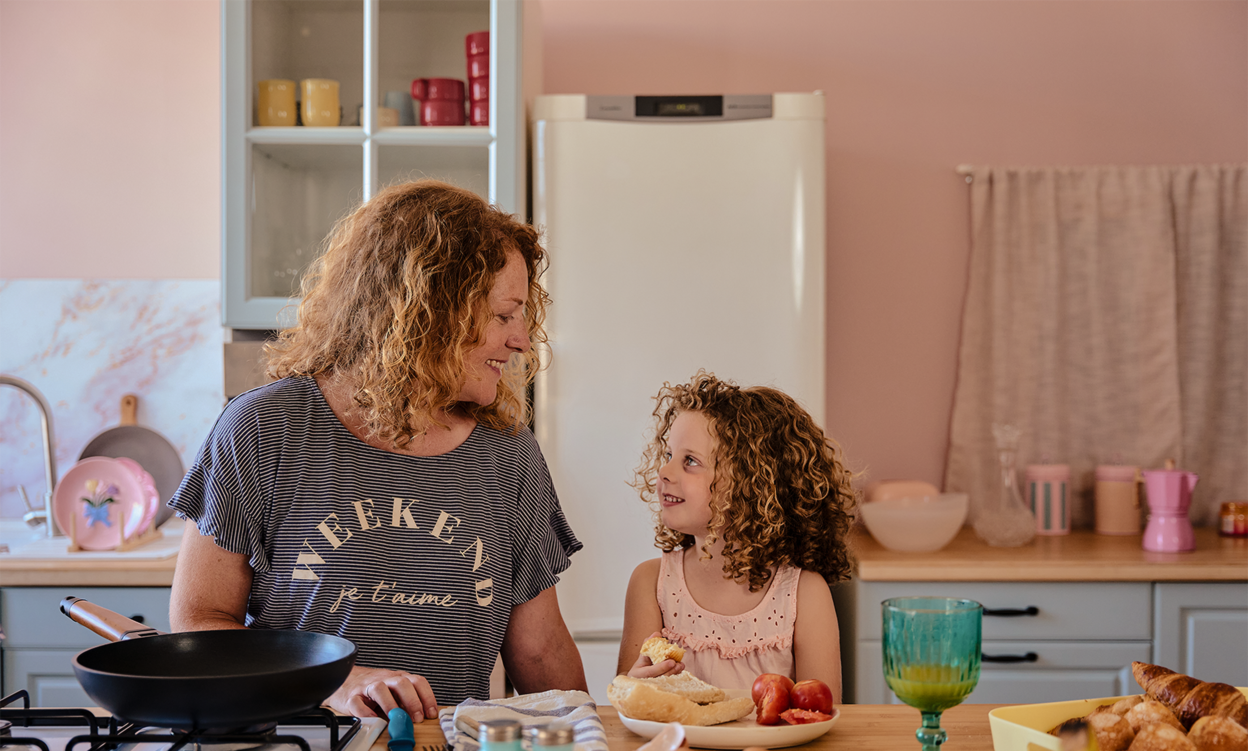 A woman and a young girl with curly hair smiling and talking in a kitchen, surrounded by breakfast foods and kitchen items.